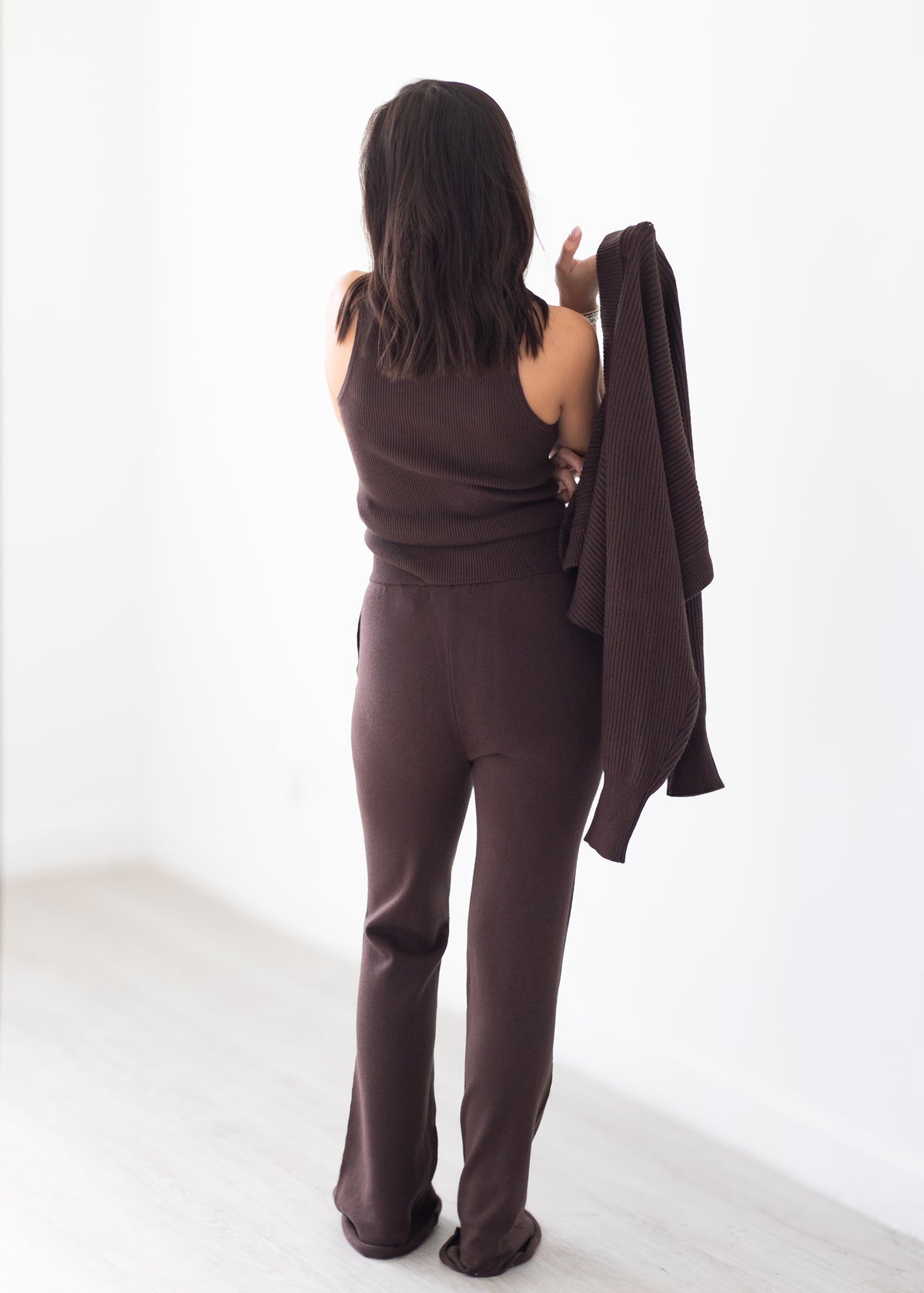 A woman stands with her back to the camera wearing a matching dark brown lounge set consisting of a sleeveless top and wide-leg pants, while holding a dark brown cardigan against a white wall.