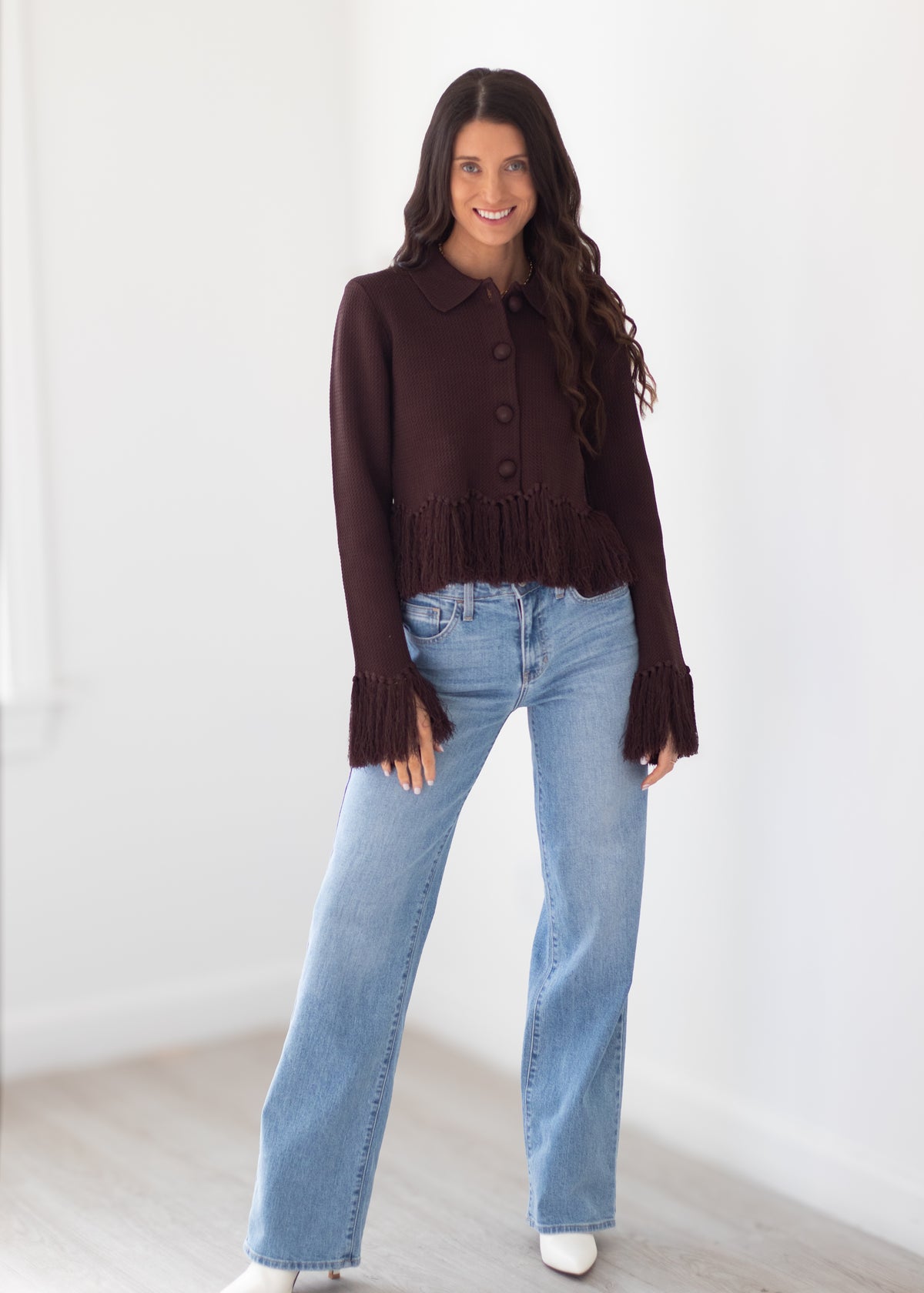 A woman modeling a dark brown cropped button-front cardigan with fringe trim and flared cuffs, paired with light-wash blue jeans, standing in a bright indoor room with white walls and light wood flooring.