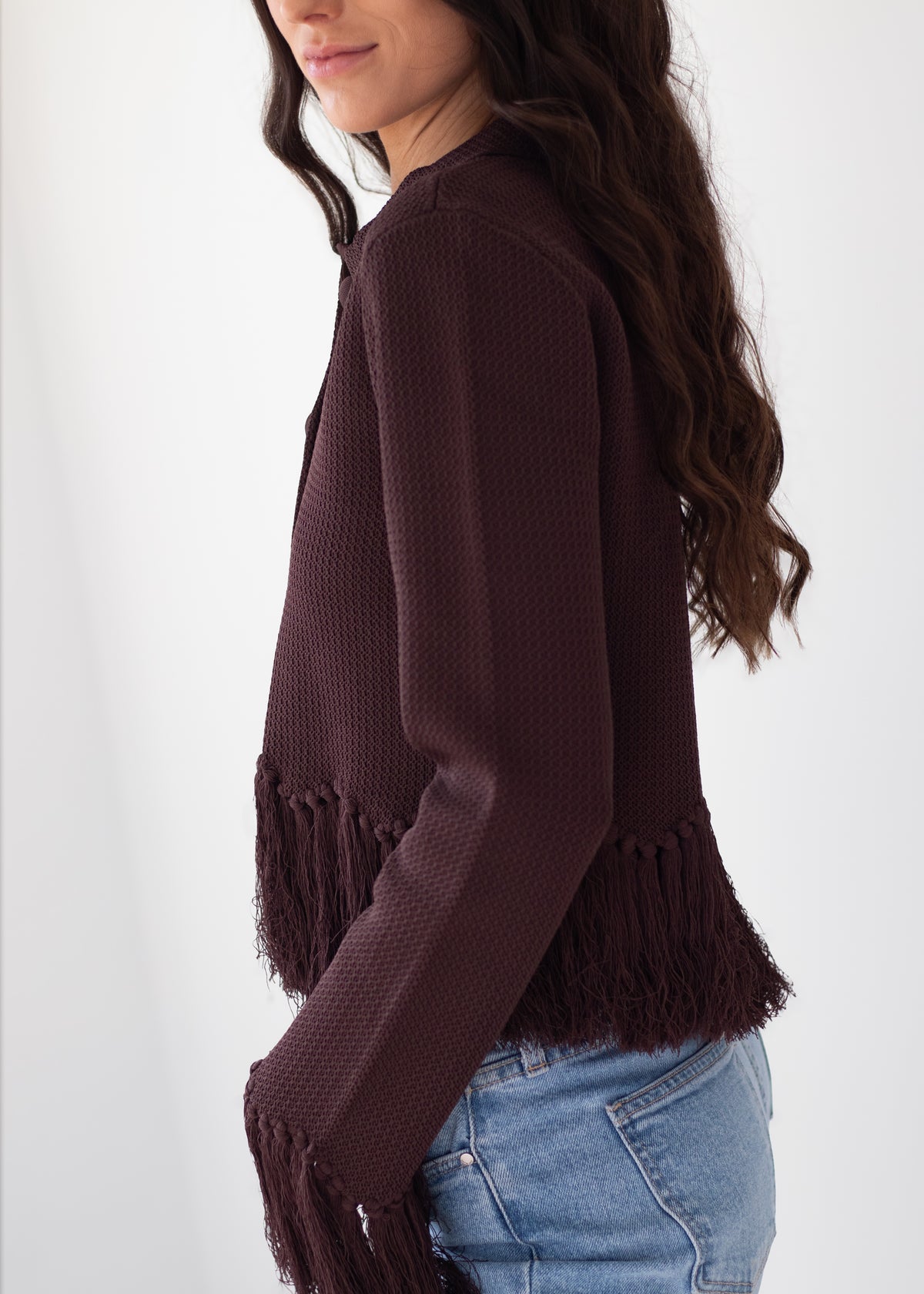 A woman wearing a dark brown waffle-knit cropped sweater with fringe detailing at the hem and cuffs, paired with light blue jeans, shown from a side profile against a plain white background.