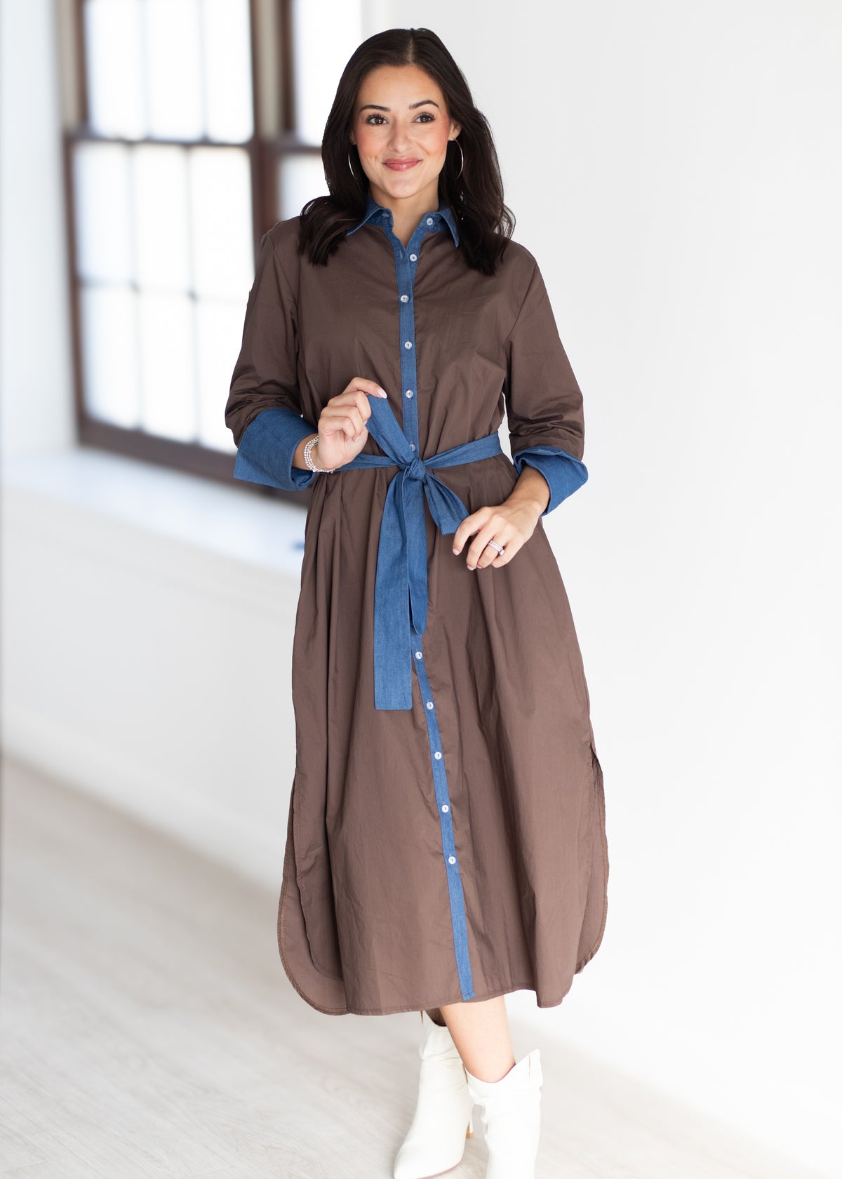 A woman modeling a brown button-front shirt dress with blue shirt-sleeves and a matching blue tie belt, paired with white ankle boots in a bright indoor room.