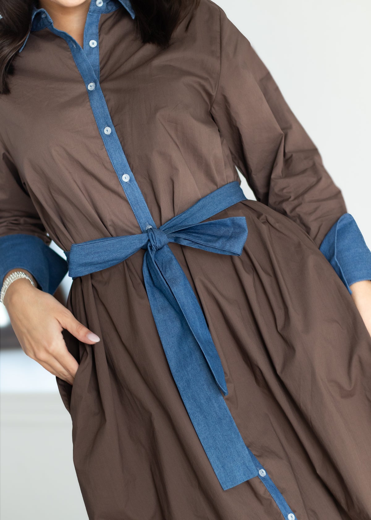 A close-up of a woman wearing a brown long-sleeve shirt dress with blue contrast trim, a blue tie belt at the waist, and blue button details down the front placket.