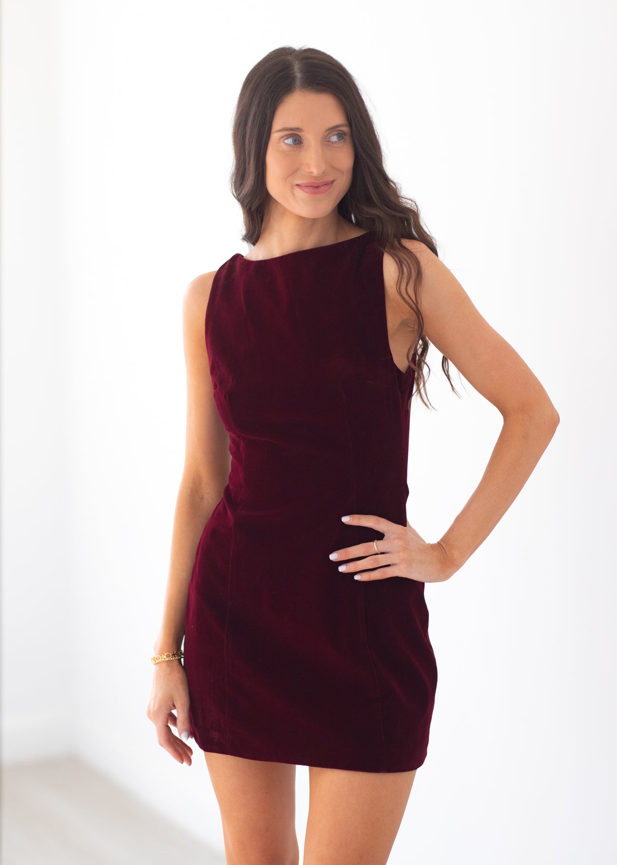 A woman modeling a sleeveless, off-the-shoulder mini dress in deep burgundy velvet against a bright white studio background.