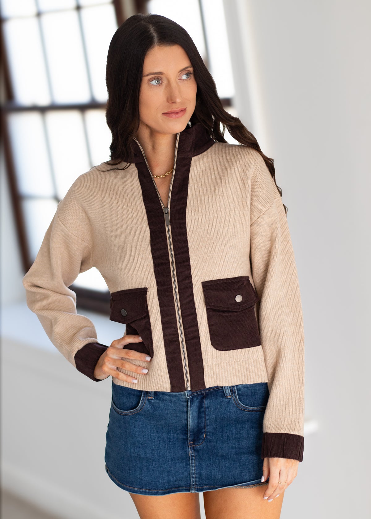 A woman modeling a beige and dark brown zip-up cardigan sweater with front pockets, paired with a blue denim mini skirt, in a bright indoor setting.