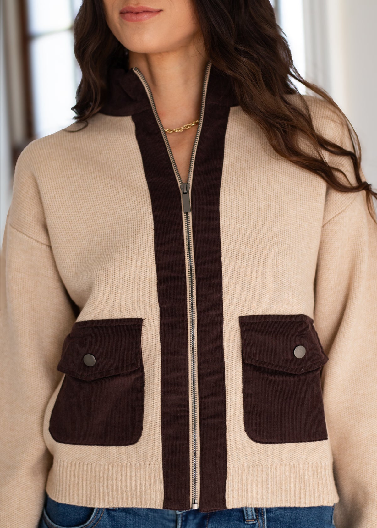 A woman wearing a beige knit zip-up cardigan with dark brown contrast panels, including a hood, zipper trim, and two front flap pockets, paired with blue jeans.