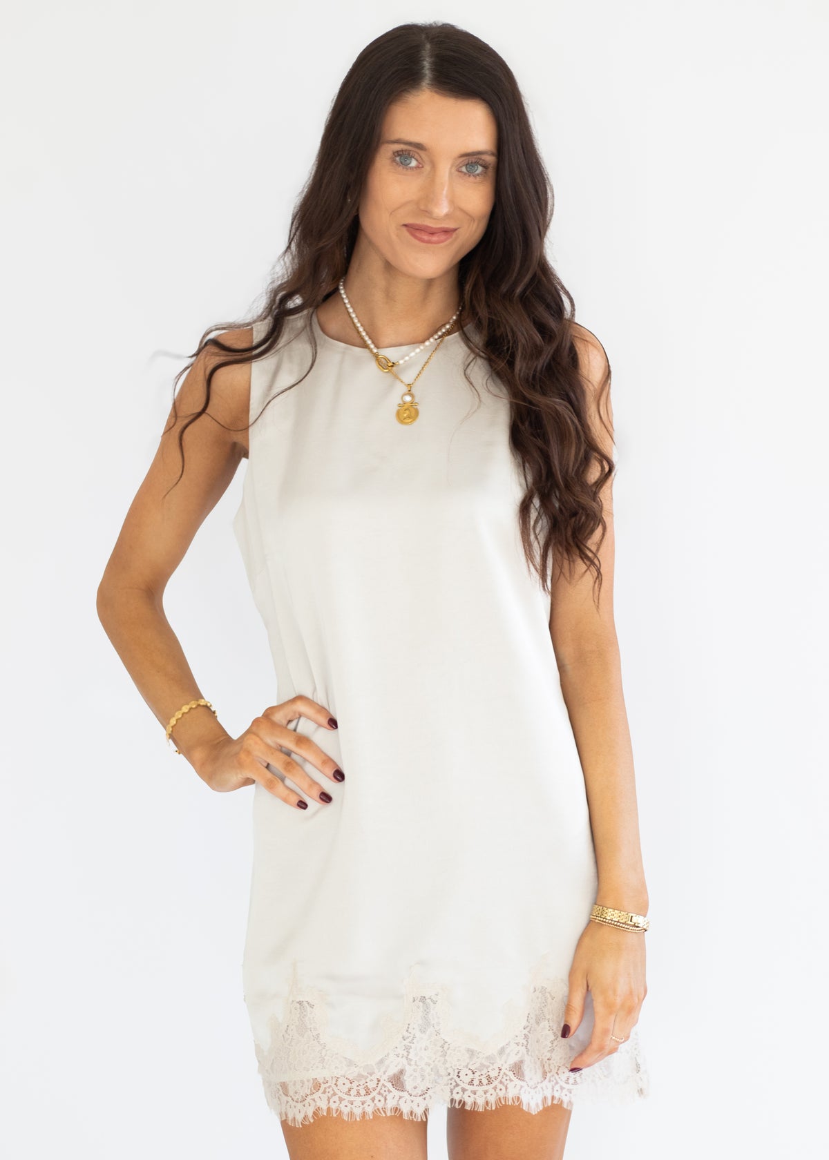 A woman modeling a sleeveless white mini dress with a delicate lace hem, styled with gold jewelry against a plain white background.