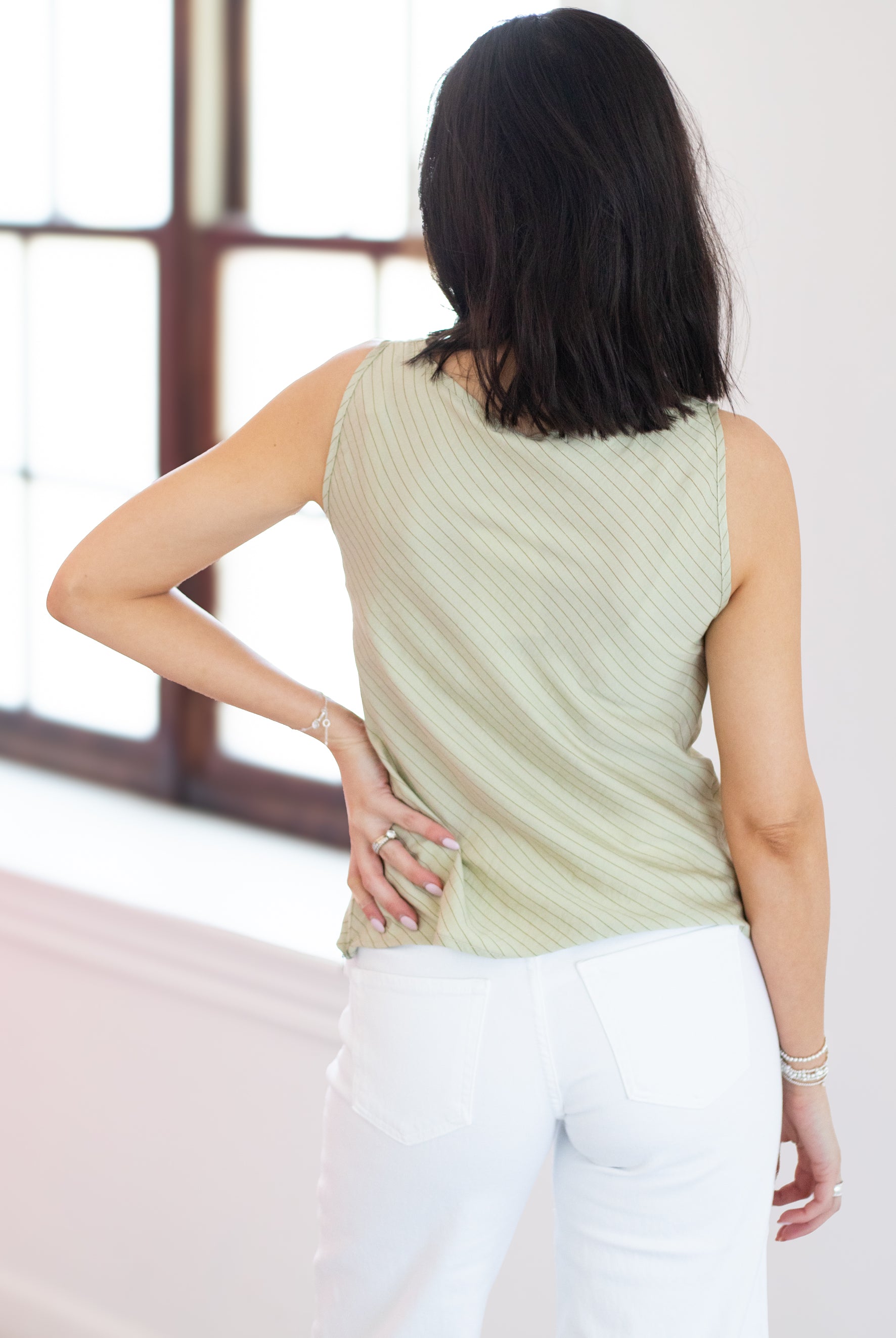 Woman wearing a light green sleeveless top and white pants, standing in a room with large windows.