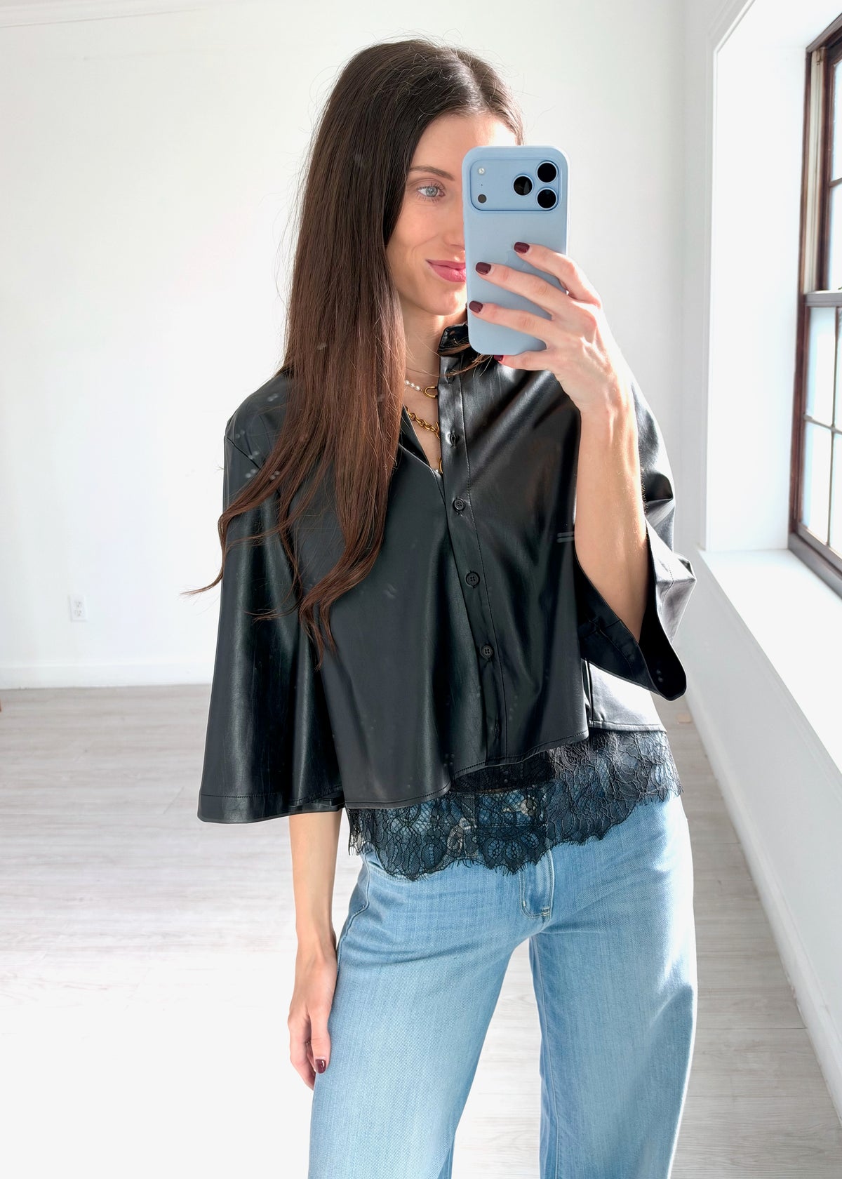 A woman takes a mirror selfie in a bright room, wearing a black faux-leather cropped button-up shirt with wide three-quarter sleeves and a black lace hem, paired with light blue jeans.