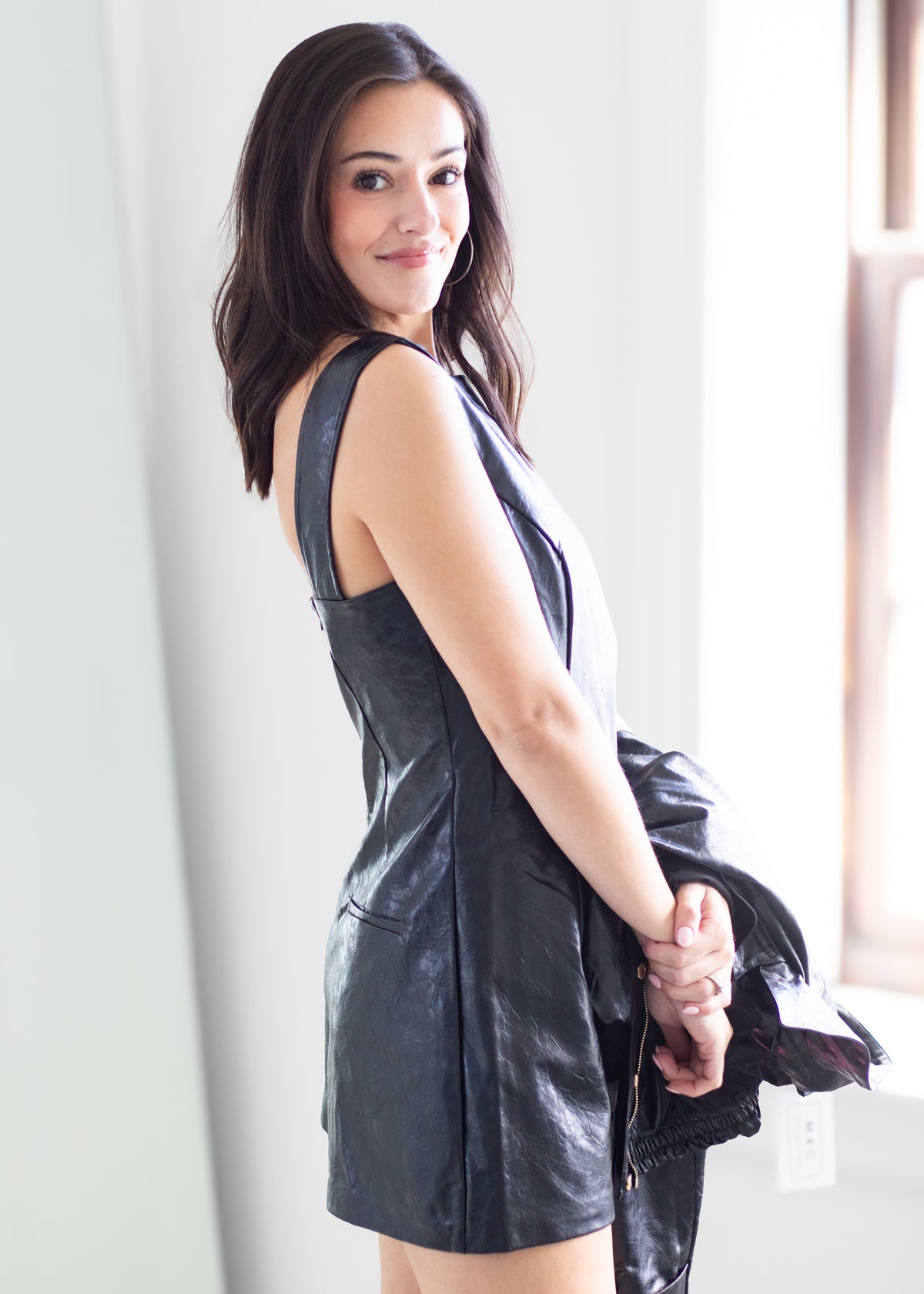A woman wearing a black faux-leather sleeveless mini dress, holding a matching black jacket in a bright indoor setting.