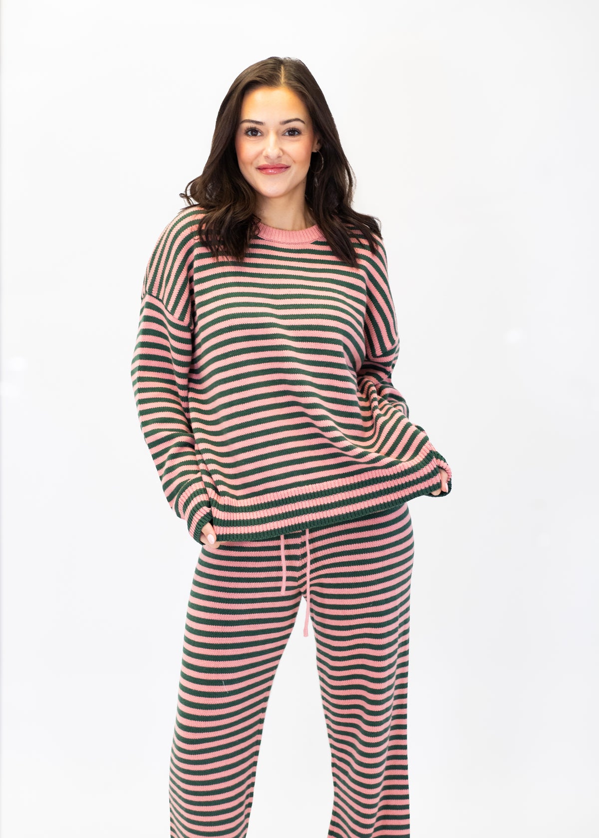 A woman modeling a matching two-piece loungewear set featuring a long-sleeve knit top and sweatpants with horizontal stripes in pink, dark green, and cream, set against a plain white background.