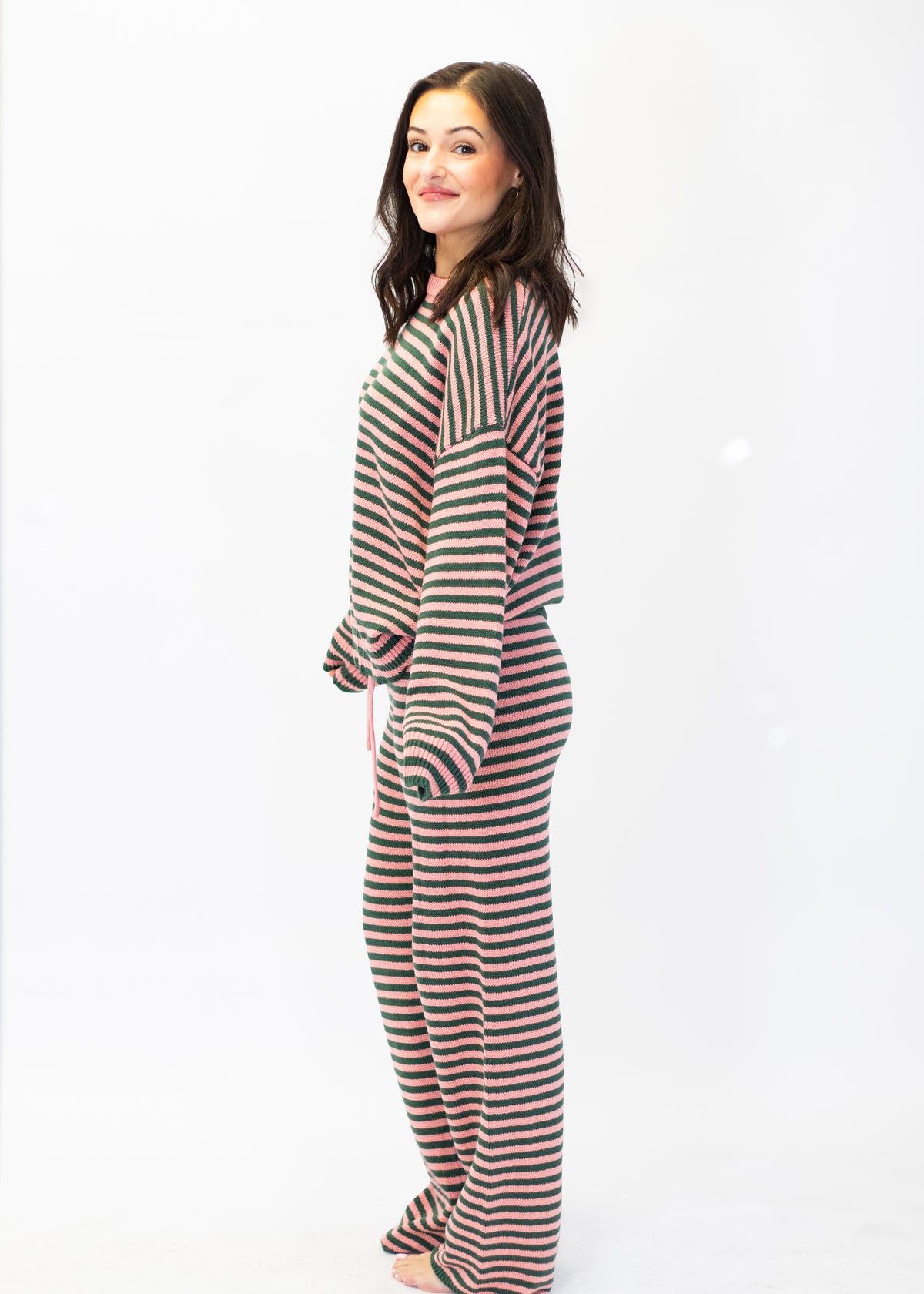 A woman modeling a matching two-piece loungewear set with horizontal stripes in dark green, red, and cream, standing against a plain white studio background.