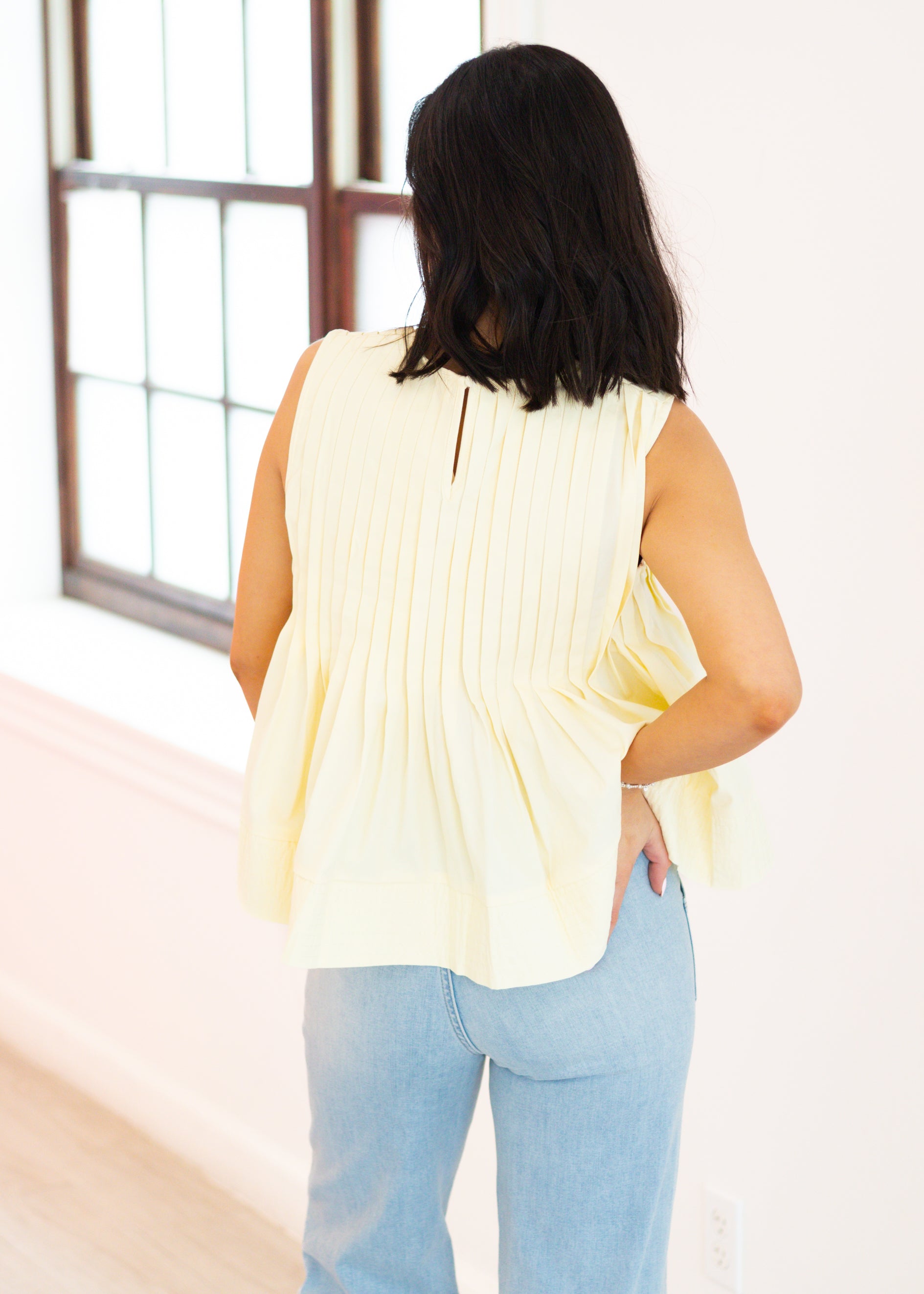 Woman wearing a light yellow sleeveless top and light blue jeans, standing in a room with a window.
