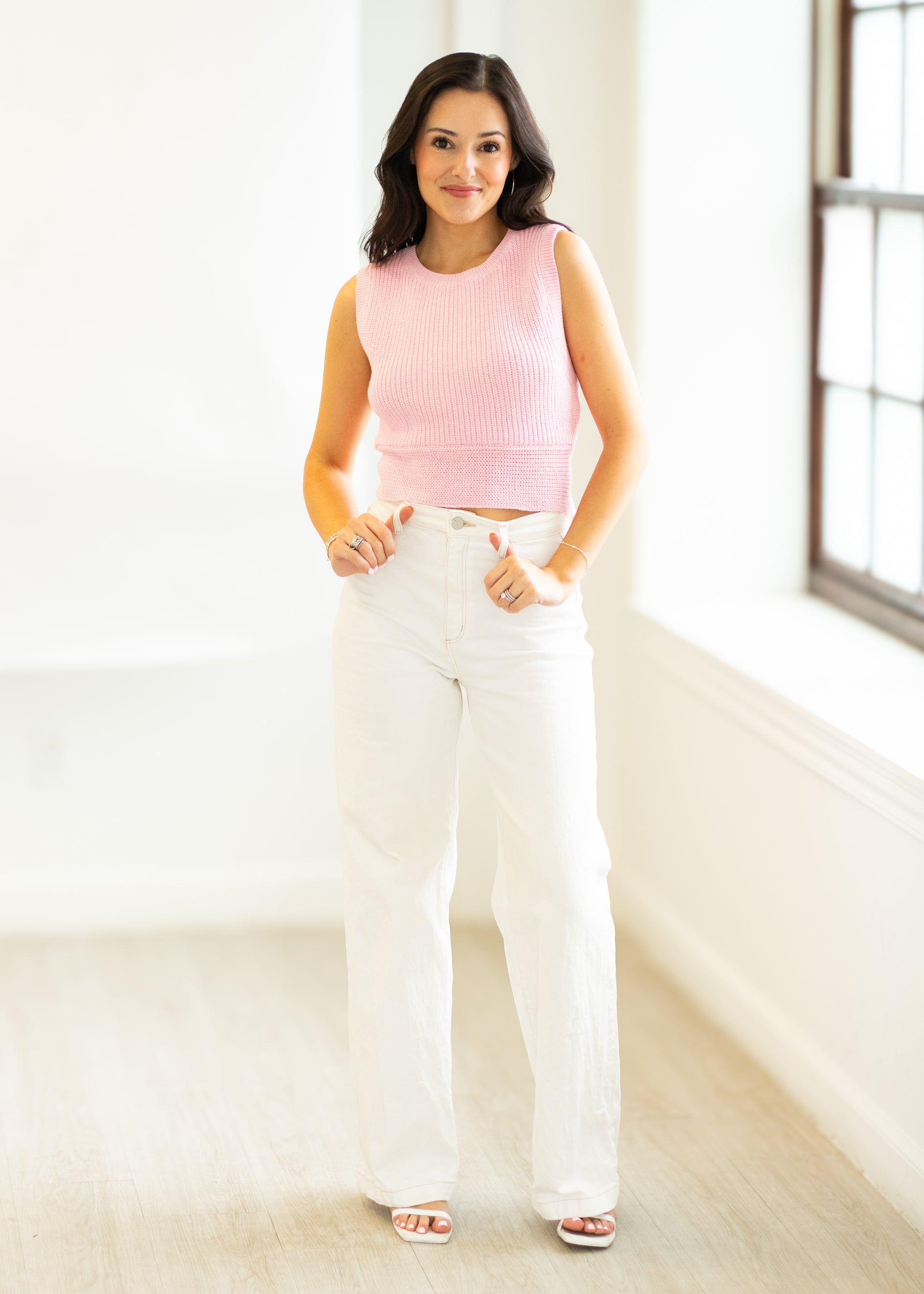 Woman wearing a pink sleeveless top and white pants in a bright room.