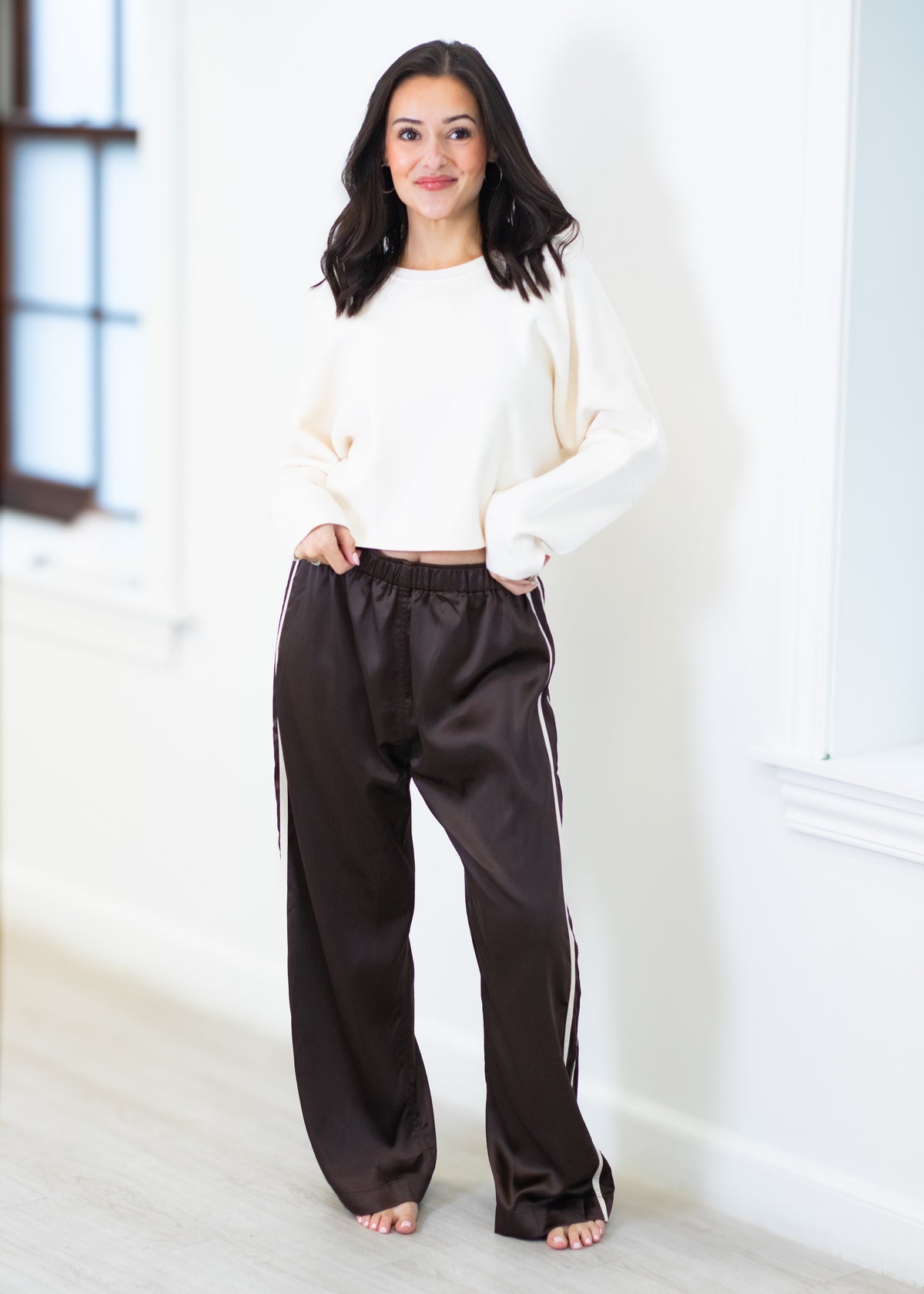 A woman stands barefoot indoors wearing a cream cropped long-sleeve top and dark brown wide-leg satin pants with white side stripes.