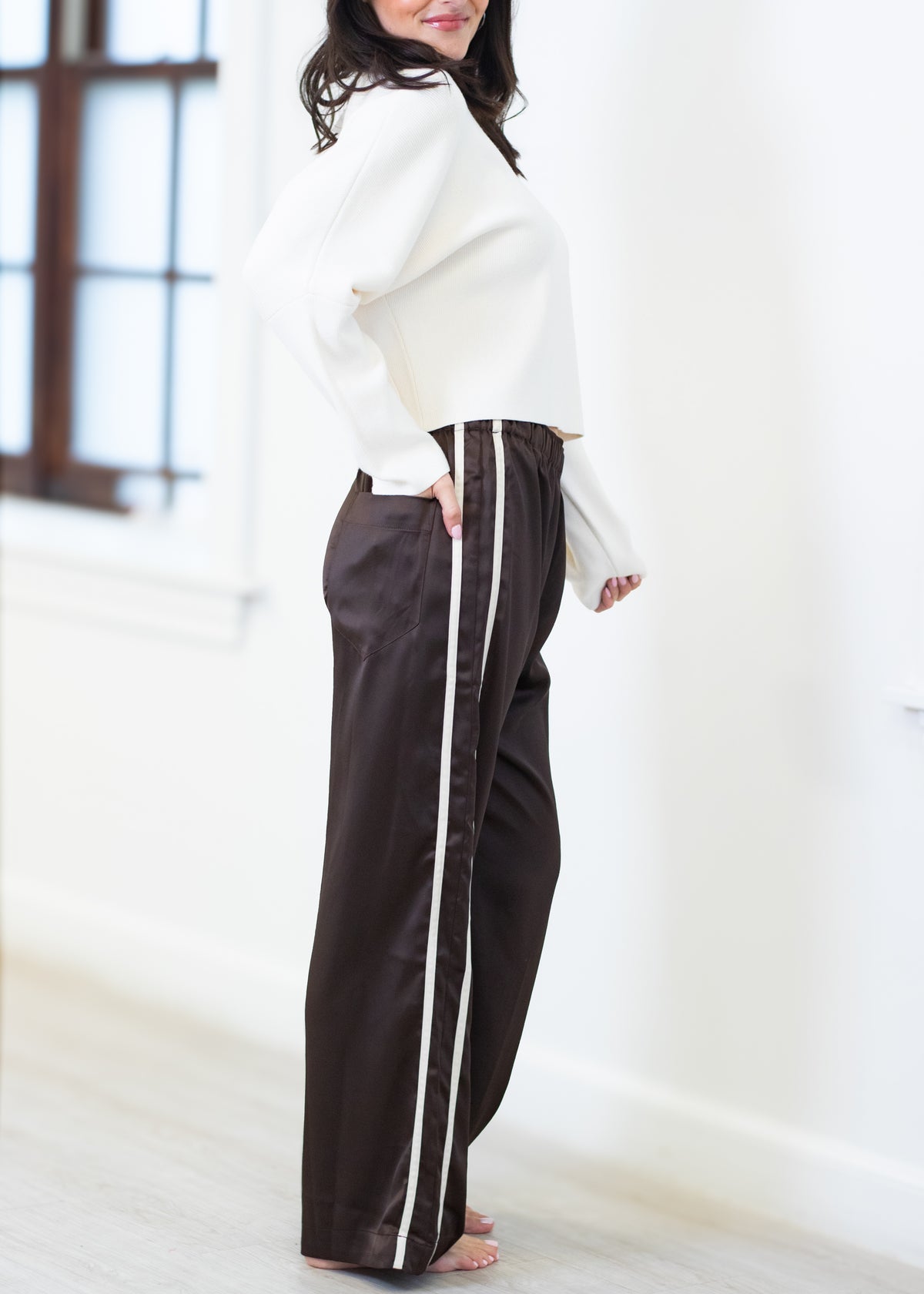 A woman wearing dark brown wide-leg pants with a white side stripe, paired with a white cropped sweater, standing in a bright room.