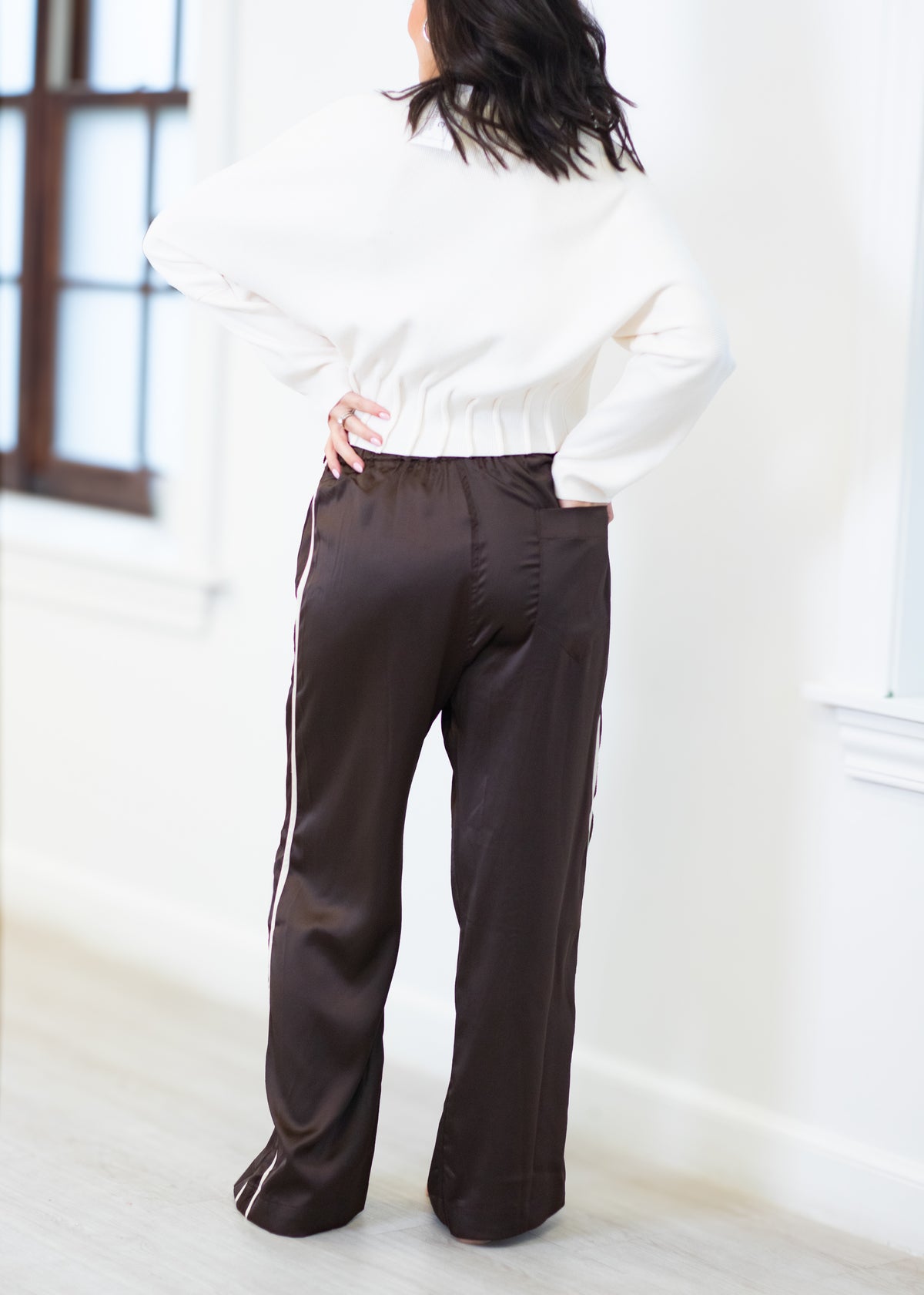 A person shown from the back wearing dark brown wide-leg satin pants with white side stripes and a white knit sweater, standing in a bright indoor room.