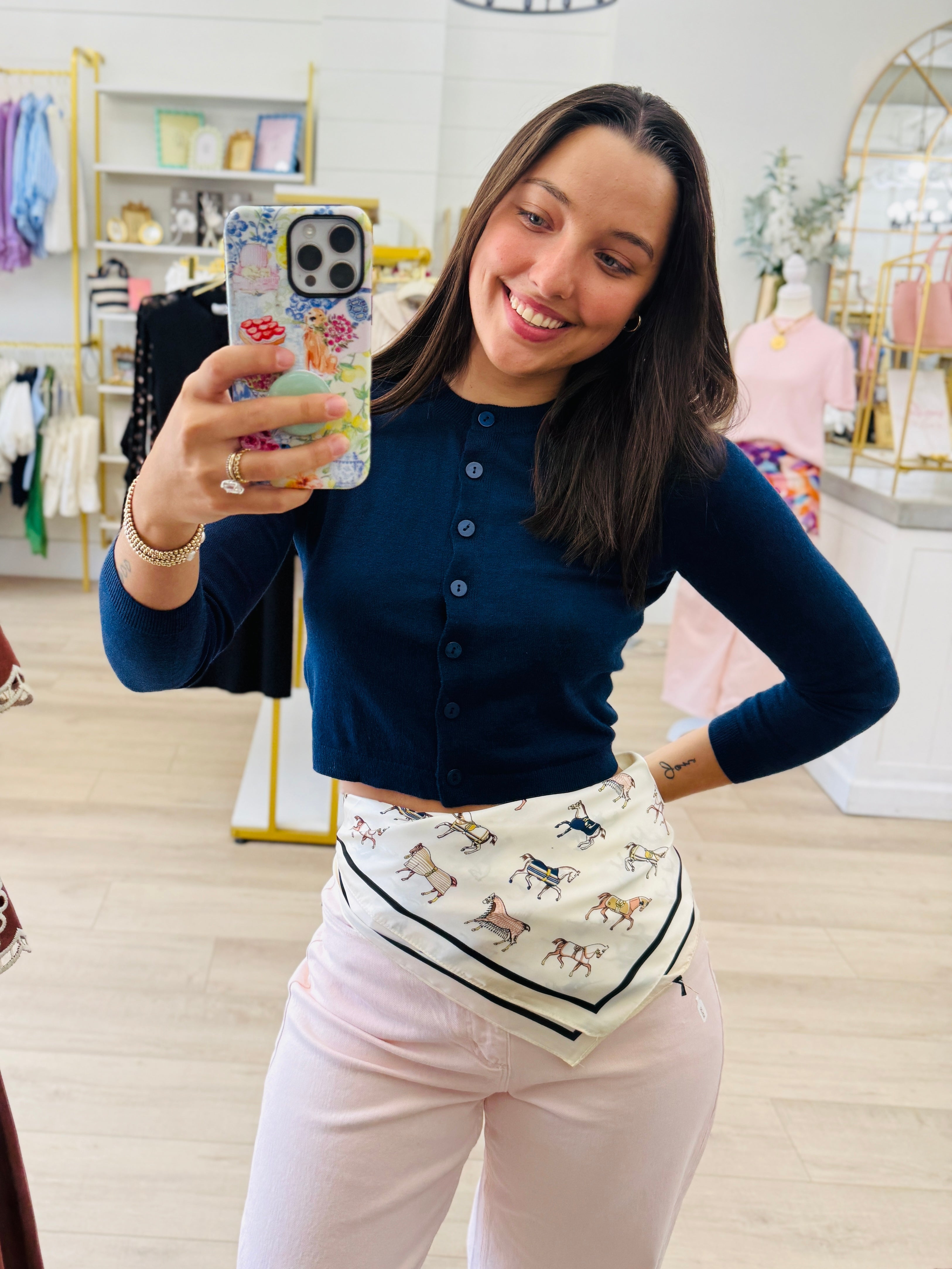 A woman takes a mirror selfie in a boutique, wearing a navy blue long-sleeve button-up top, light pink pants, and a cream scarf with a horse pattern tied around her waist.
