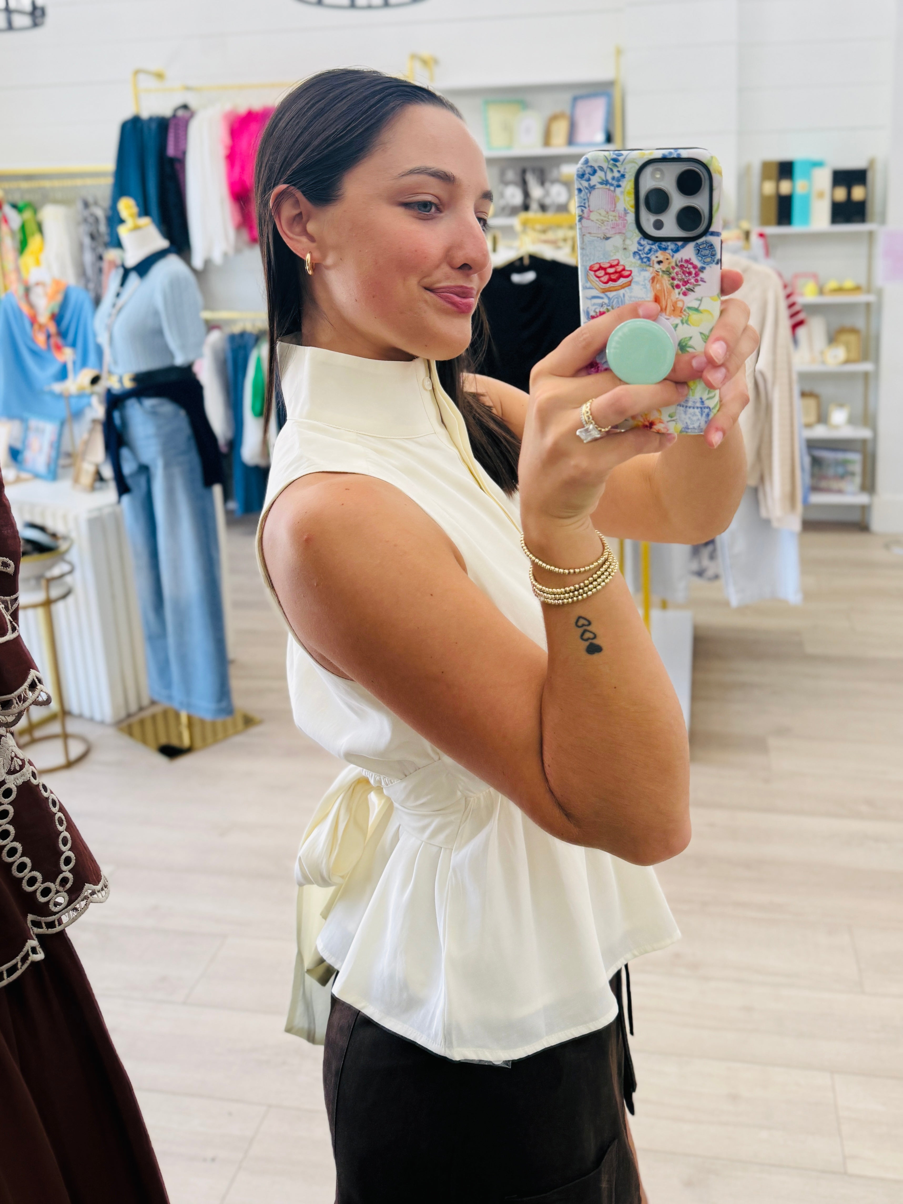 A woman taking a mirror selfie in a clothing boutique, wearing a sleeveless cream-colored peplum top with a high mock neck and a side tie, paired with black pants.