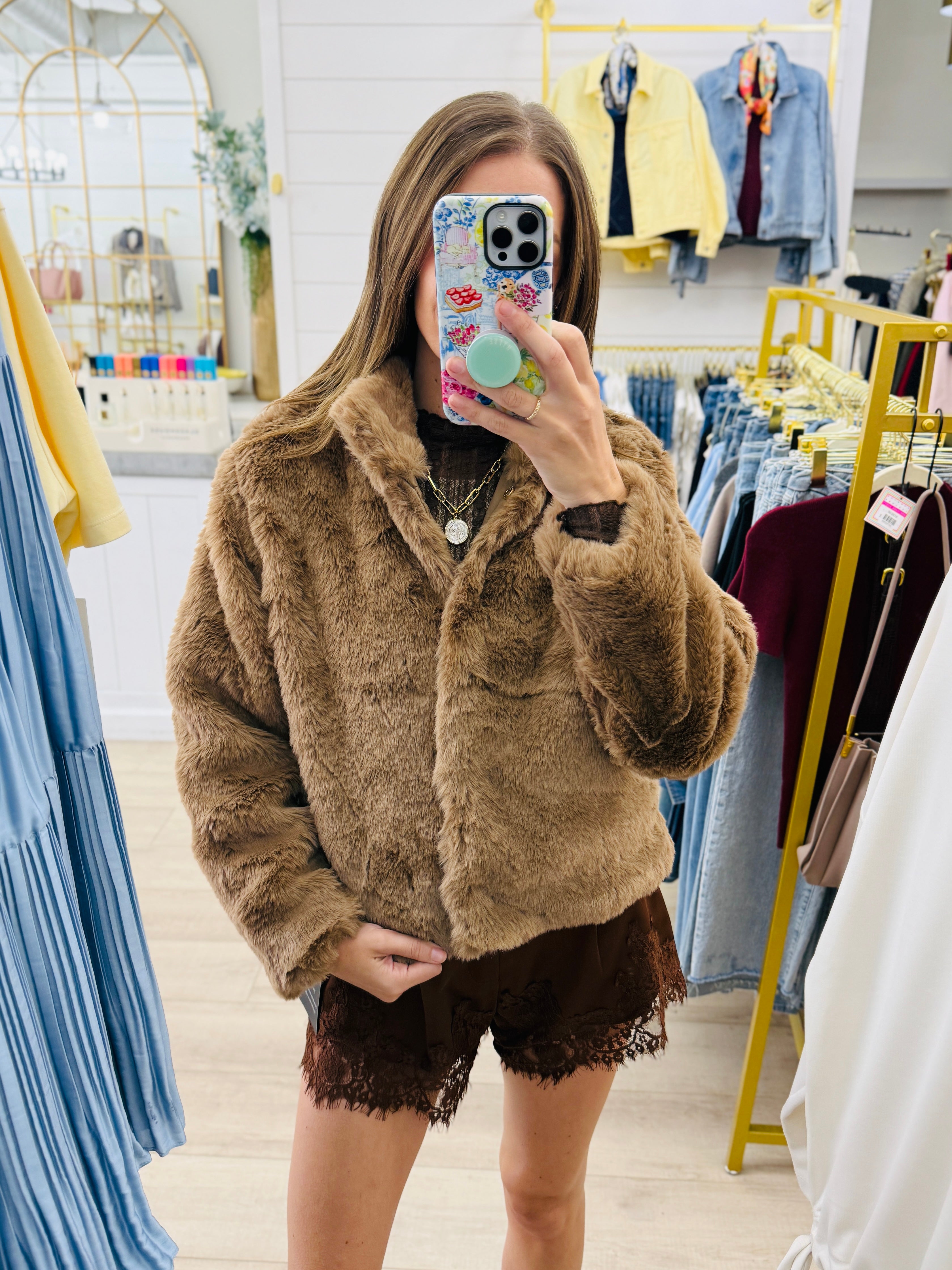 A woman takes a mirror selfie in a clothing boutique wearing a brown fuzzy faux fur jacket and brown lace-up shorts.