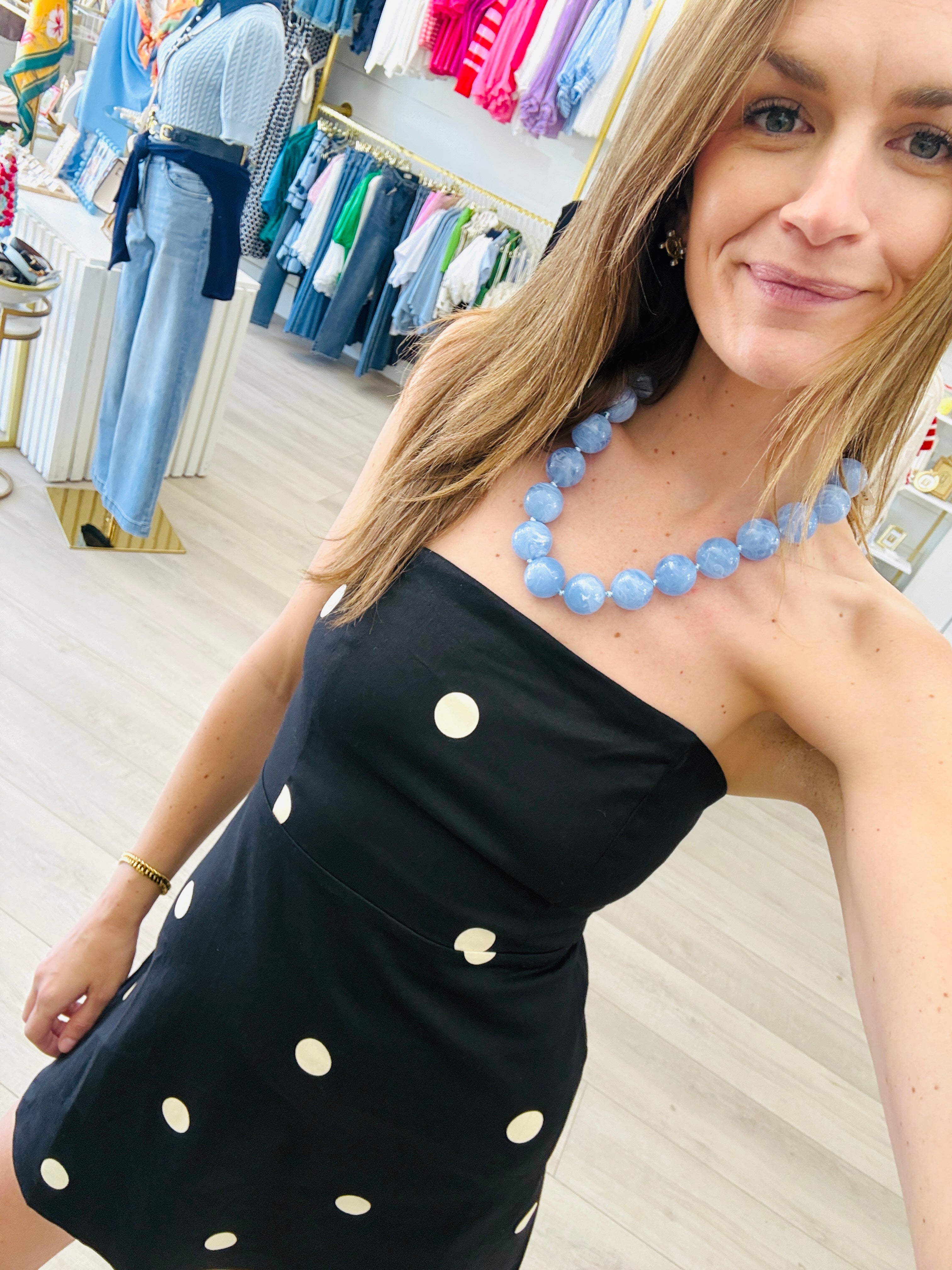A woman wearing a black strapless dress with white polka dots and a chunky light blue beaded necklace, posing inside a clothing store.