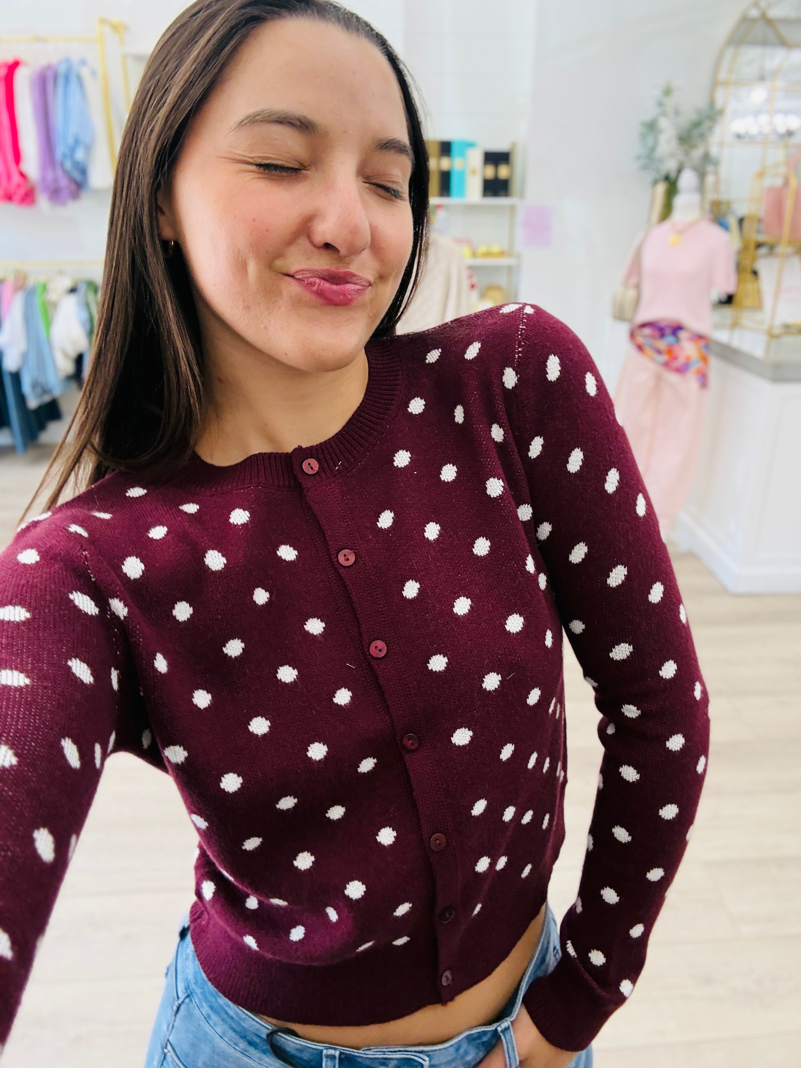A woman wearing a burgundy cardigan with white polka dots and buttons, paired with blue jeans, in a brightly lit boutique setting.
