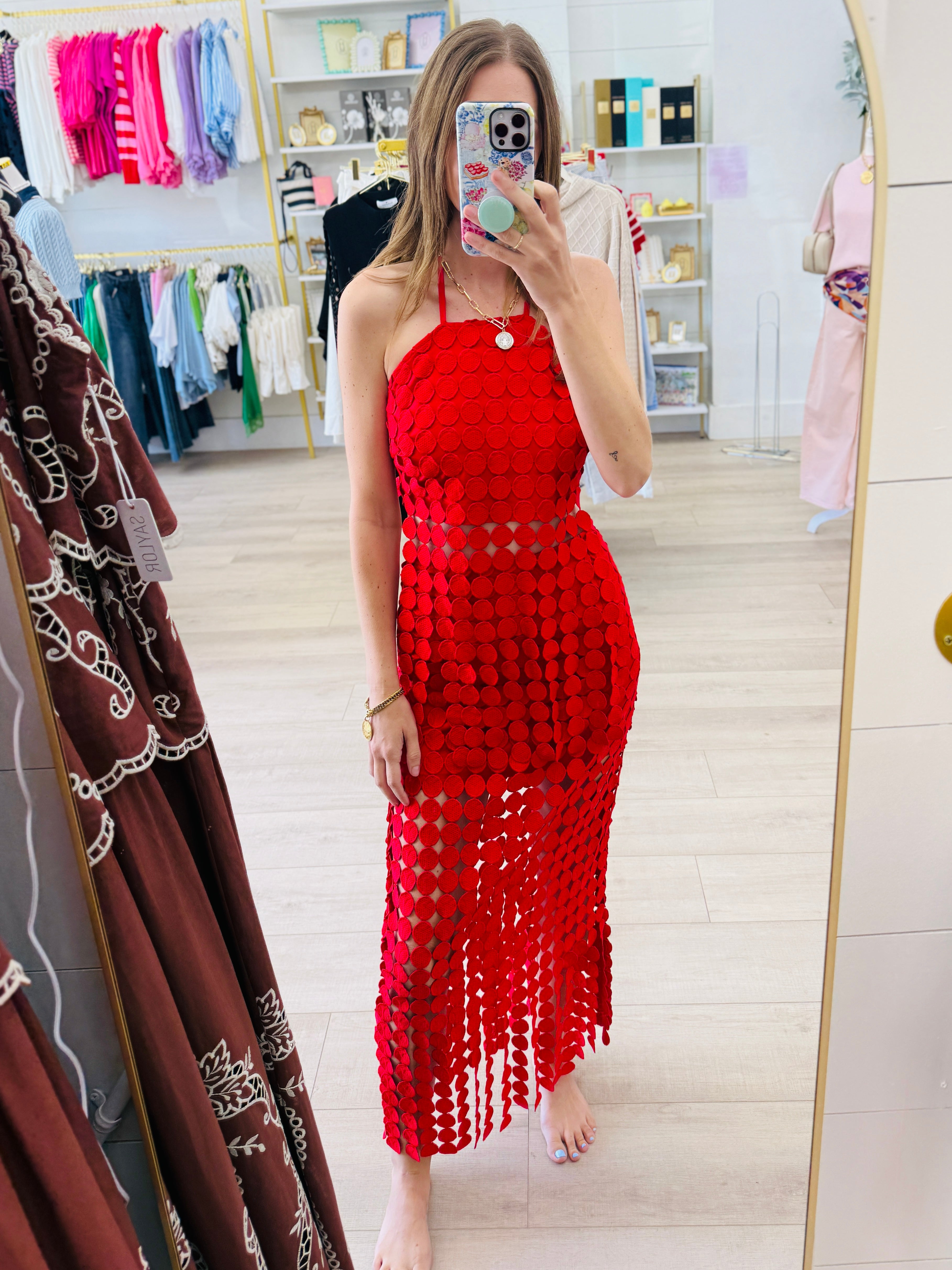 A woman takes a mirror selfie in a clothing boutique wearing a bright red crochet halter dress with a fringe hem.