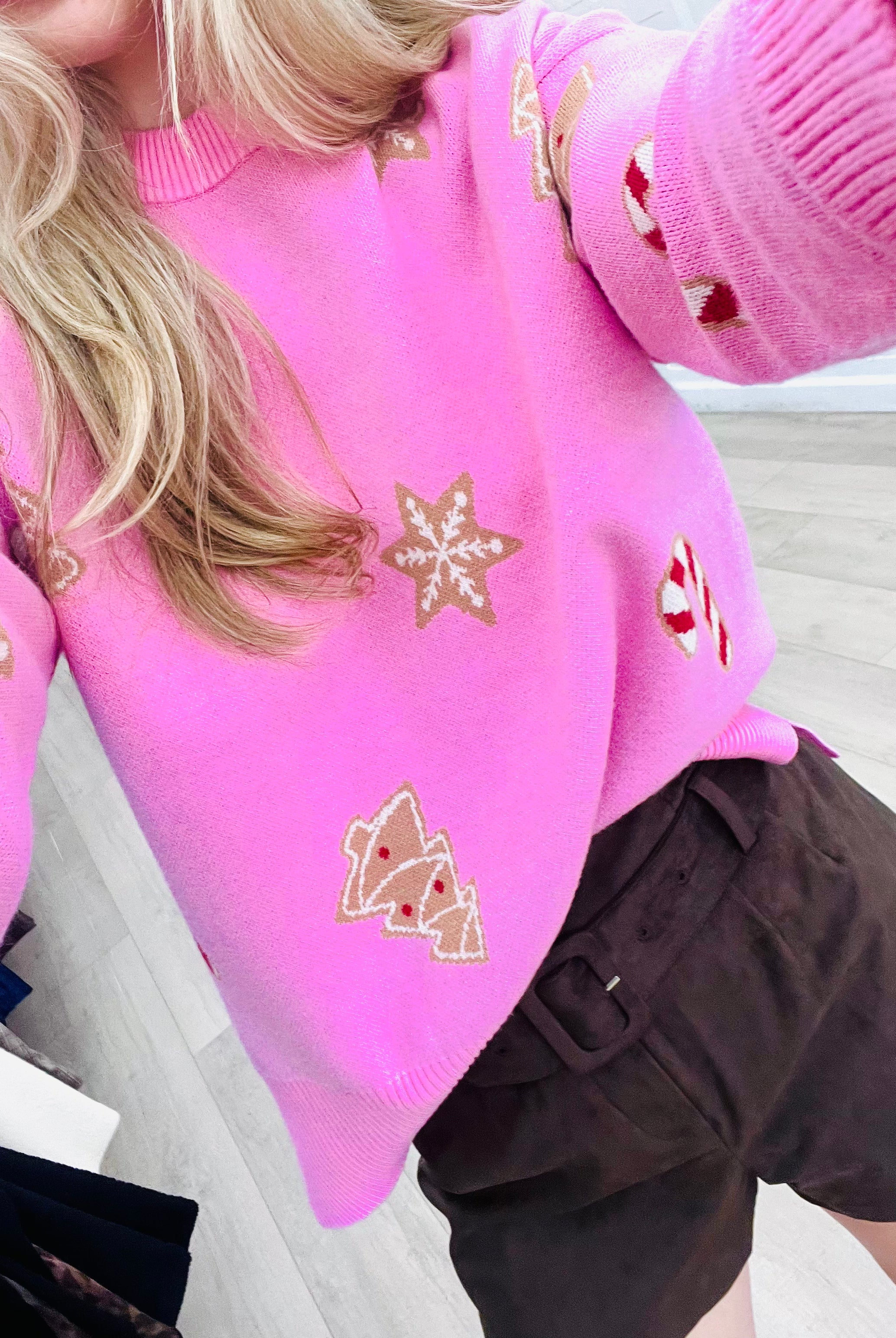 A close-up of a person wearing a bright pink knit sweater with embroidered gingerbread motifs, paired with dark brown shorts, in an indoor setting.