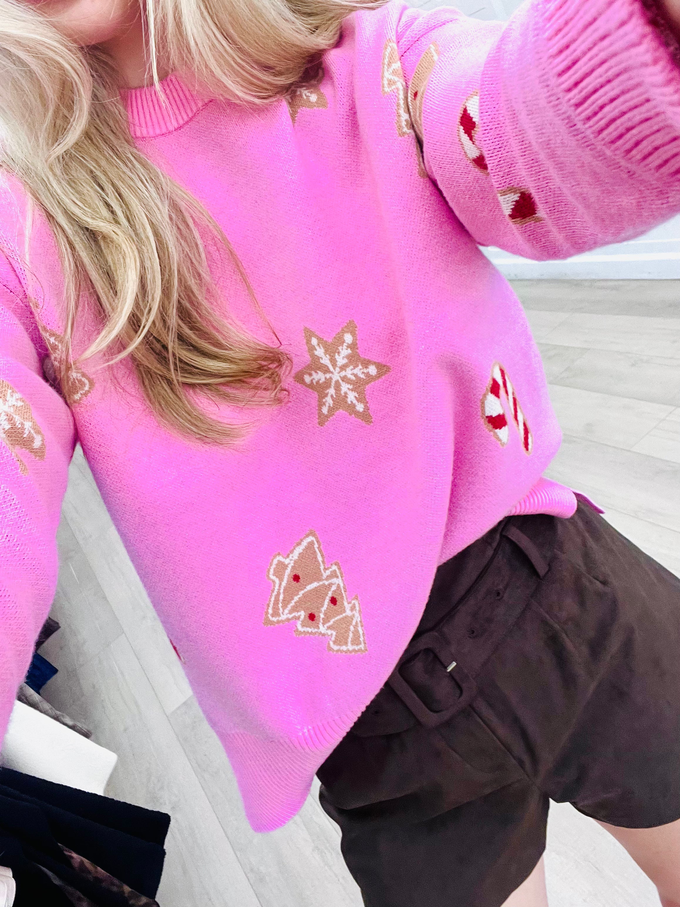 A close-up of a person wearing a bright pink knit sweater with embroidered gingerbread motifs, paired with dark brown shorts, in an indoor setting.