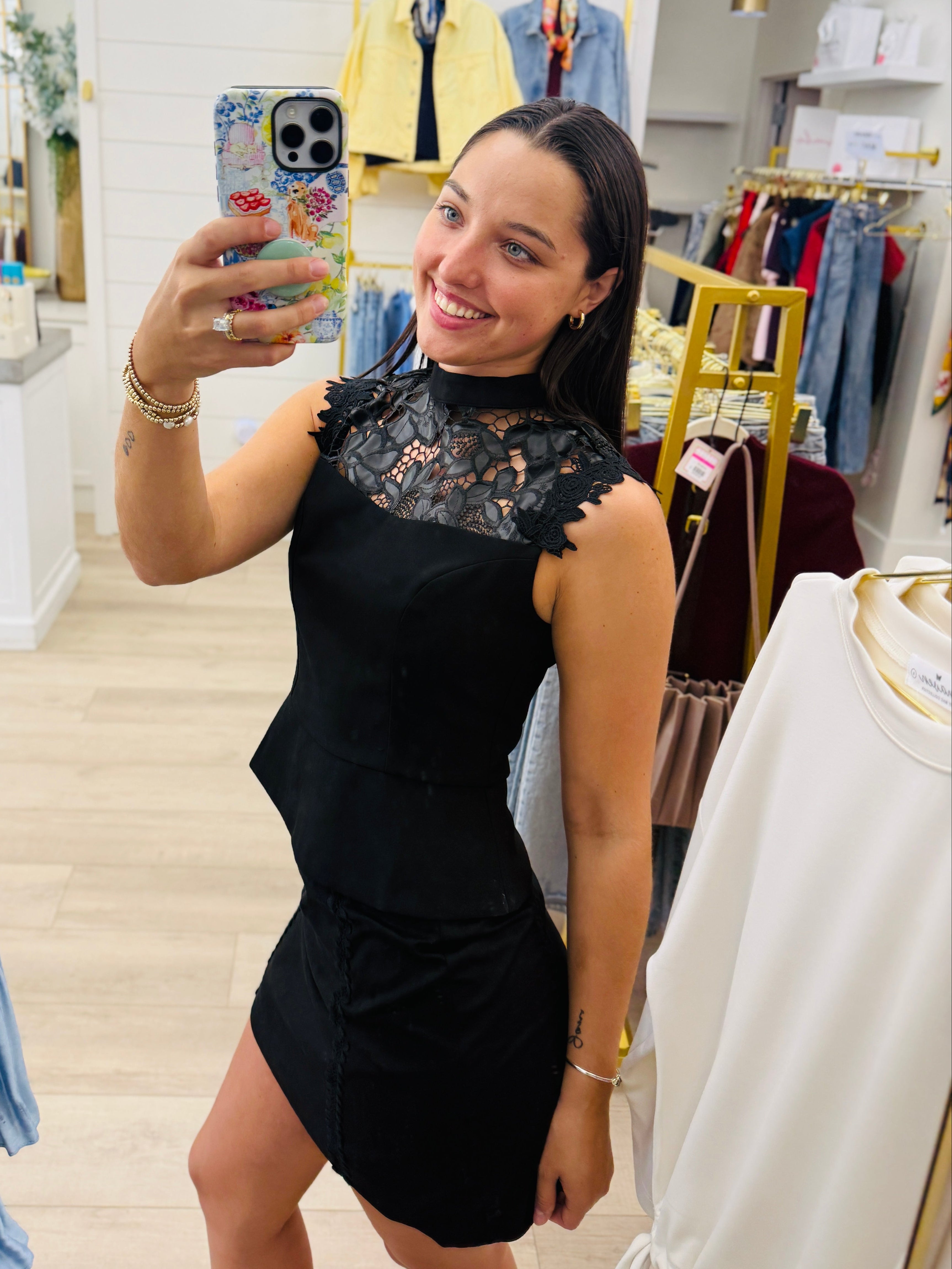 A woman takes a mirror selfie in a boutique while wearing a black sleeveless mini dress with a sheer floral lace high-neck bodice. The boutique has light wood floors, white shiplap walls, and clothing racks in the background.