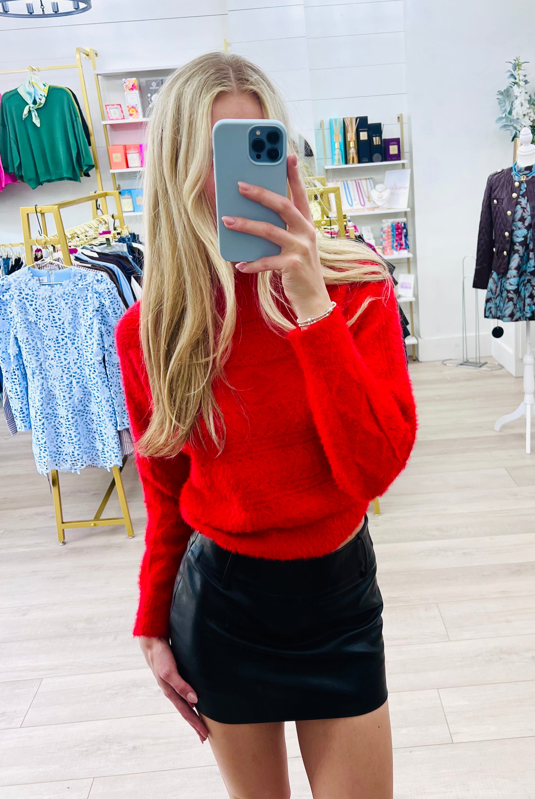 A woman takes a mirror selfie in a clothing boutique, wearing a bright red fuzzy knit sweater and a black faux-leather mini skirt.