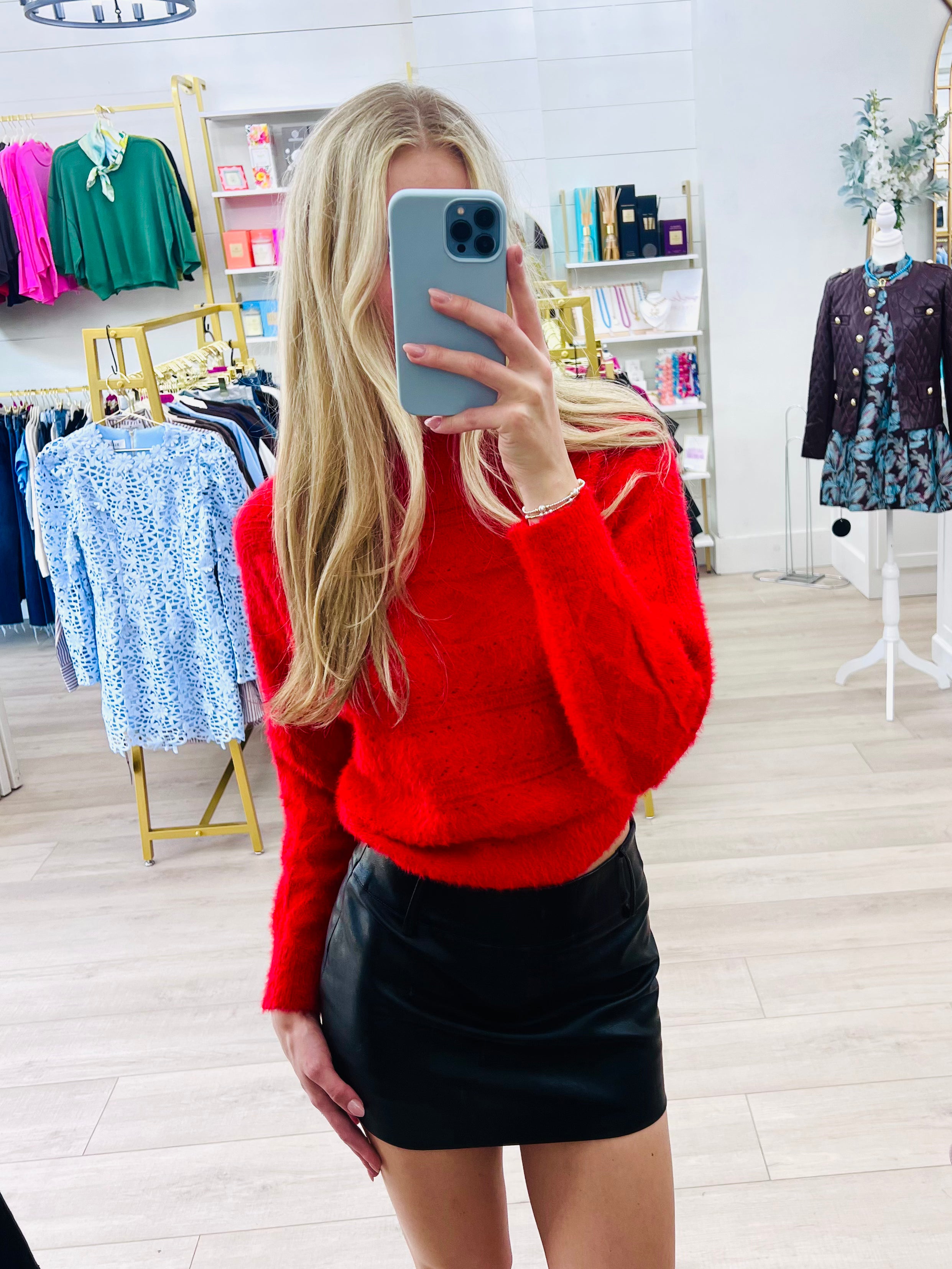 A woman takes a mirror selfie in a clothing boutique, wearing a bright red fuzzy knit sweater and a black faux-leather mini skirt.