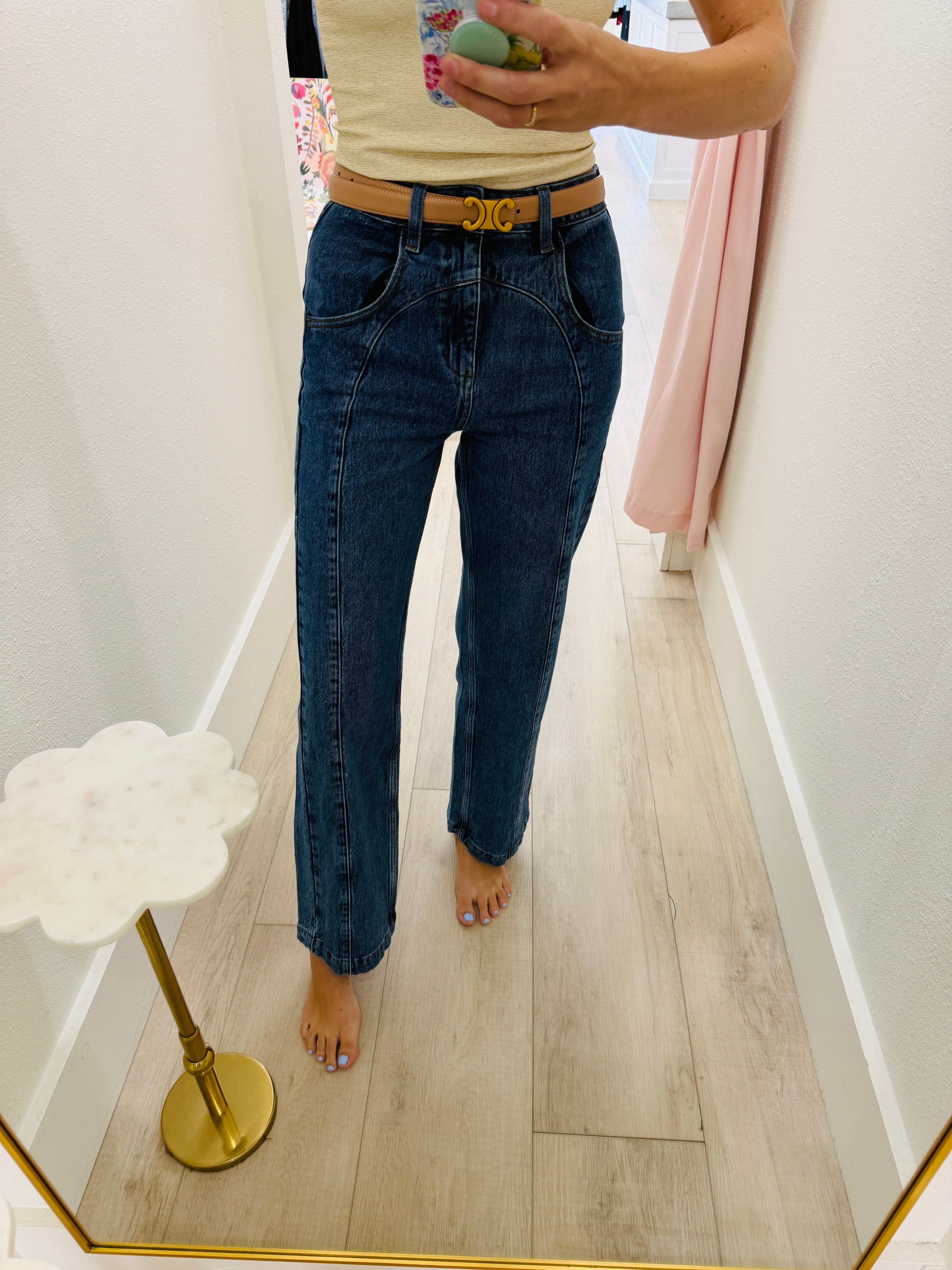 A person taking a mirror selfie in a hallway wearing dark blue straight-leg jeans, a tan belt with a gold buckle, and a light beige top.