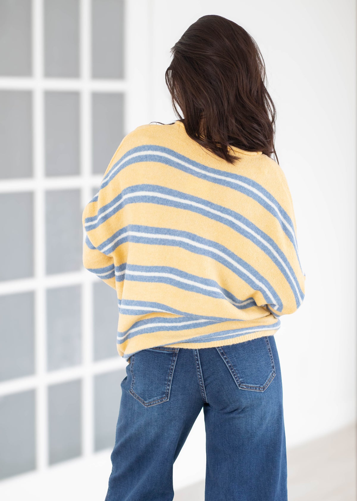 A woman viewed from behind wearing a yellow knit sweater with blue and white horizontal stripes, paired with blue jeans, standing in front of a white wall with a window grid.