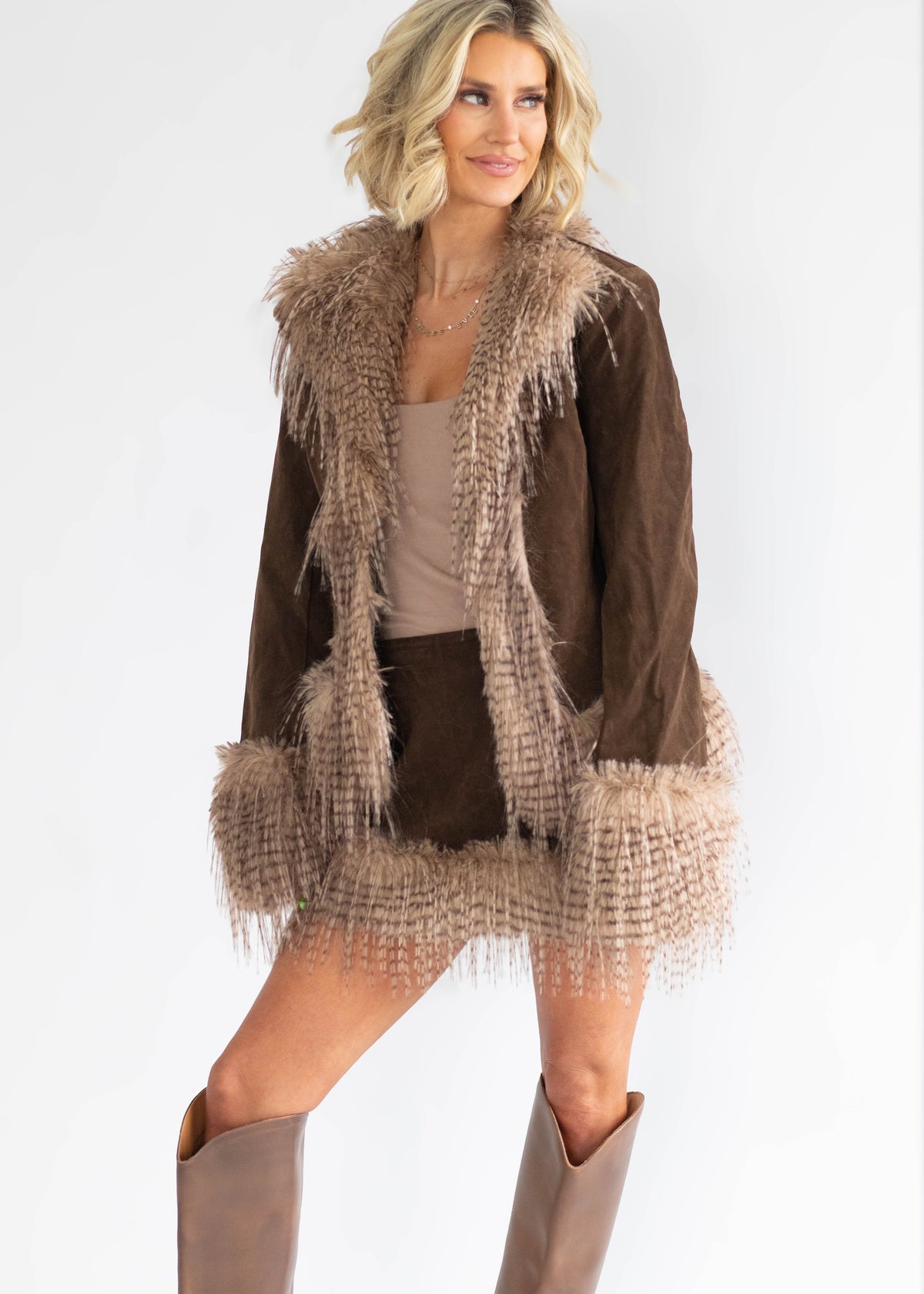 A woman modeling a brown suede jacket with beige faux fur trim, styled with a beige top, matching shorts, and beige cowboy boots against a plain white background.