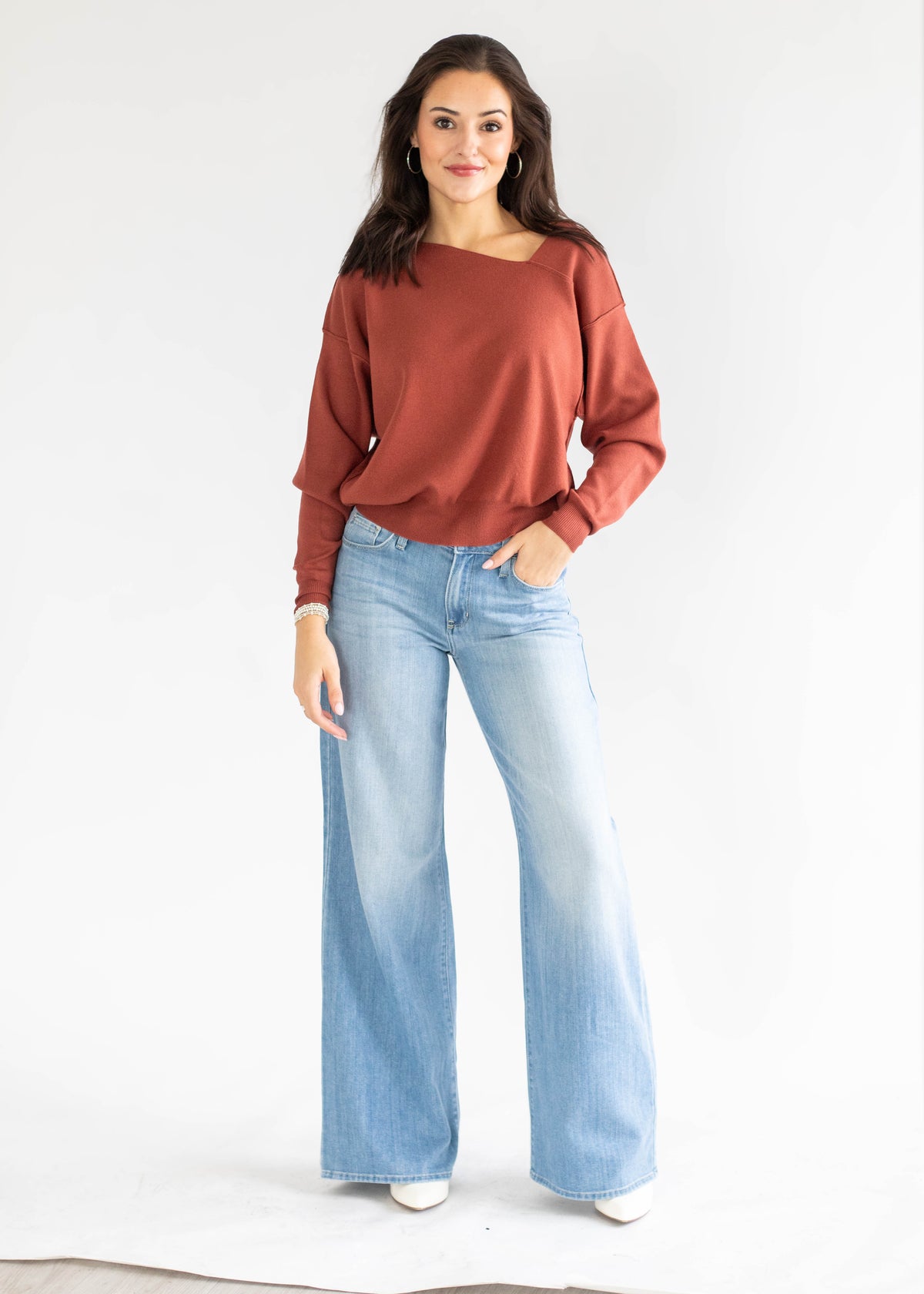A woman modeling a rust-colored, off-the-shoulder long-sleeve top paired with light-wash wide-leg jeans and white shoes, standing against a plain white studio background.
