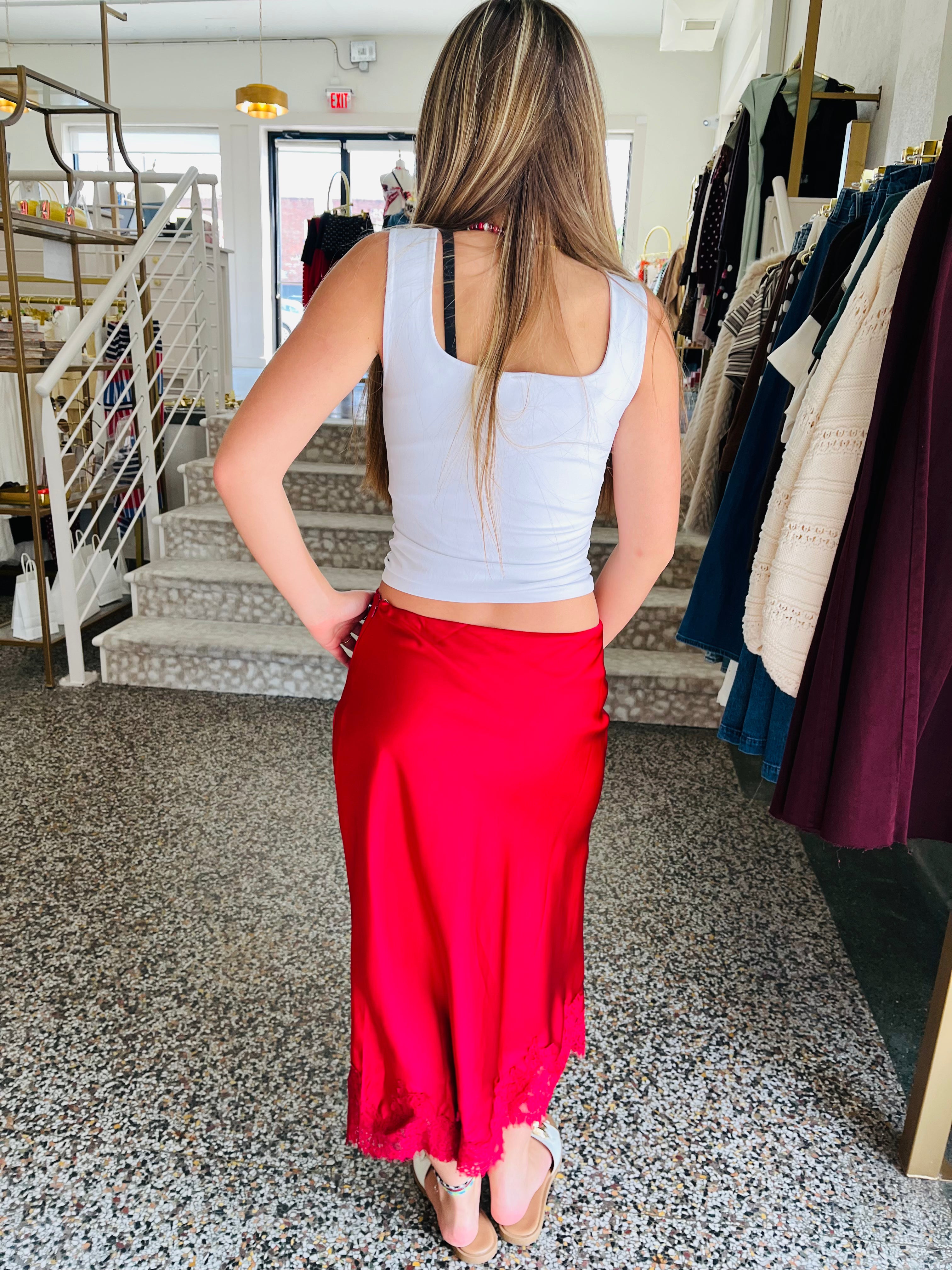 A woman standing in a boutique, viewed from behind, wearing a light gray ribbed tank top and a bright red satin maxi skirt.