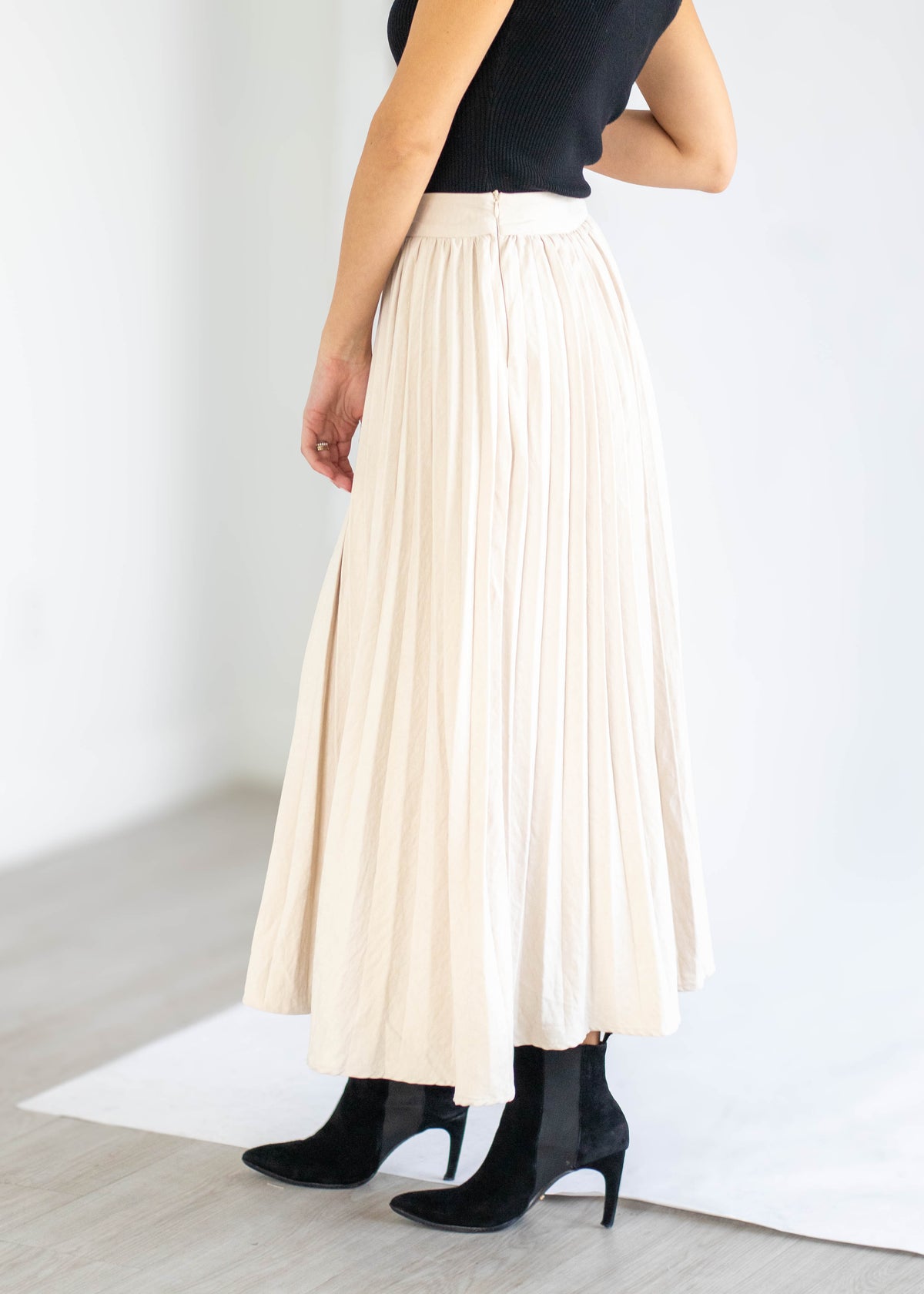 A model wearing a high-waisted, cream-colored pleated midi skirt paired with a black sleeveless top and black ankle boots, standing in a minimalist white room.