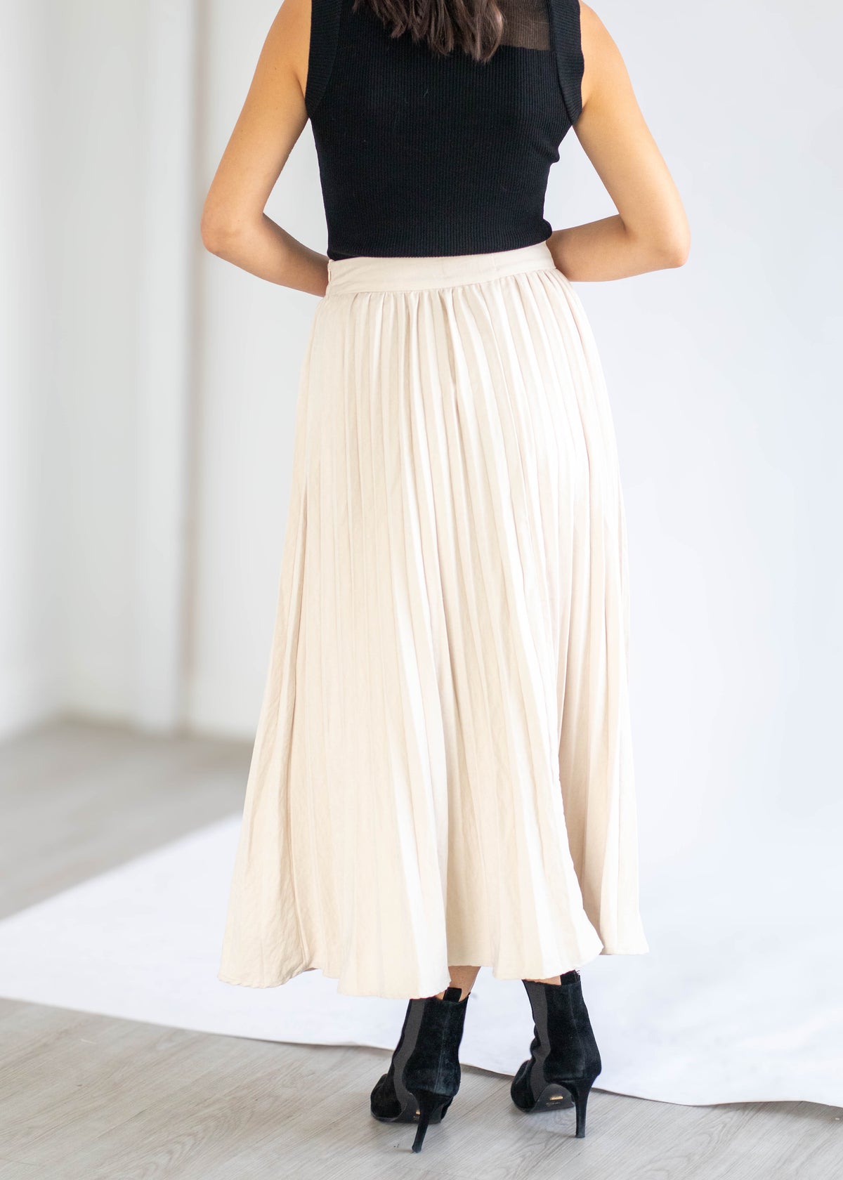 A woman shown from the back wearing a long pleated cream maxi skirt, a black sleeveless top, and black ankle boots in a bright indoor setting.