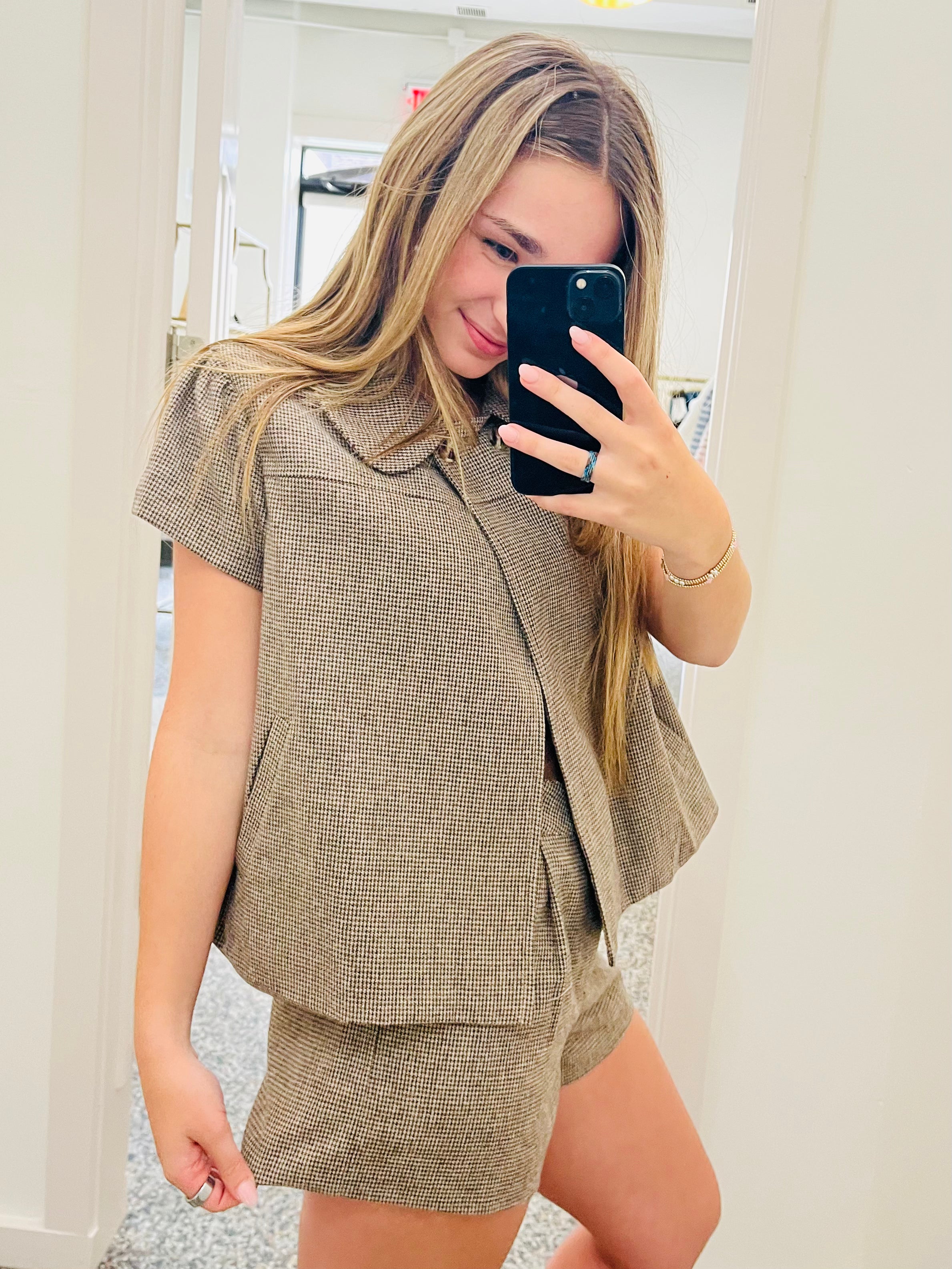 A woman takes a mirror selfie showing a matching two-piece outfit consisting of a short-sleeve button-front blazer with a large collar and matching shorts, both in a textured taupe-brown fabric.