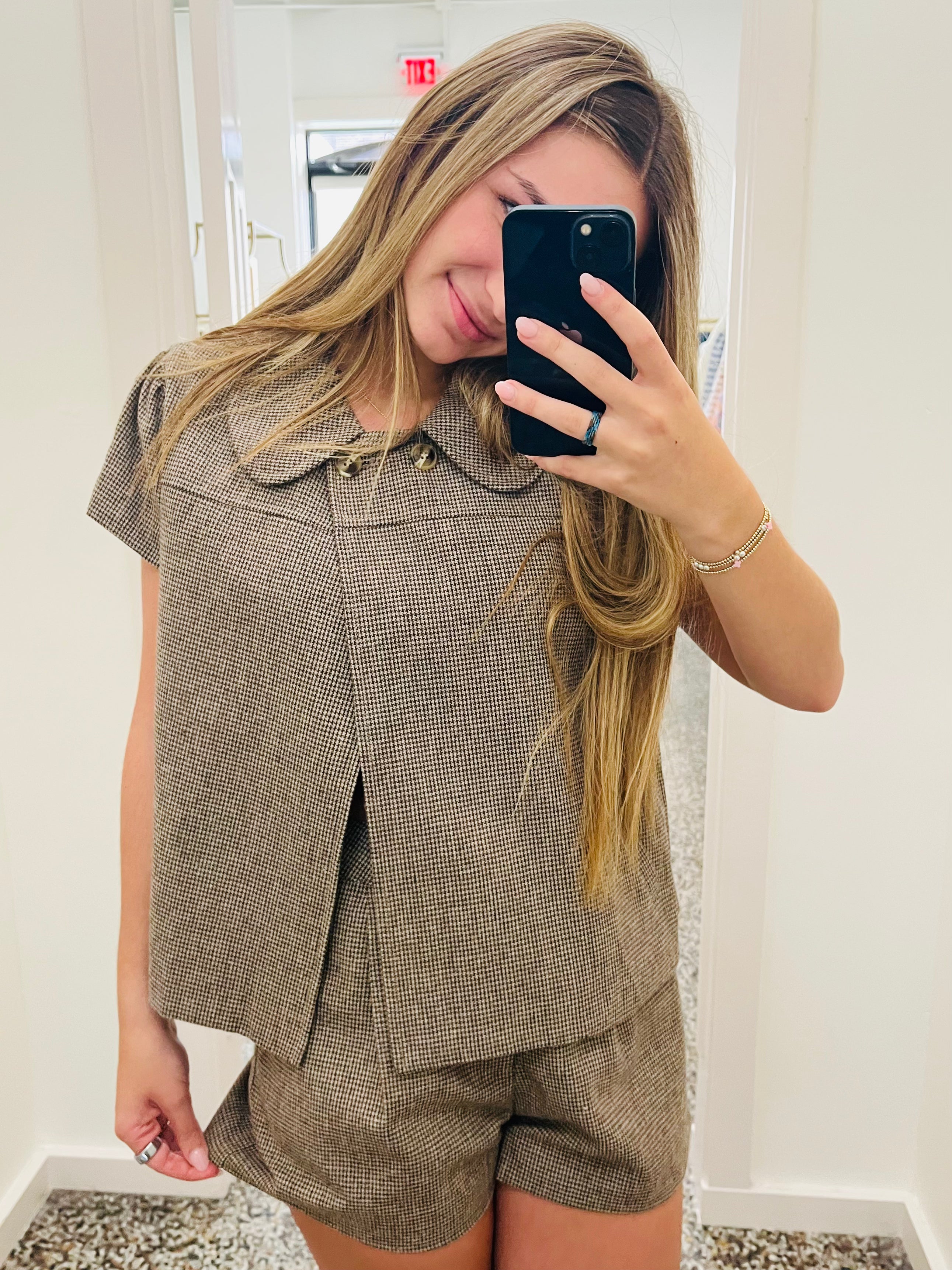 A woman takes a mirror selfie wearing a matching brown and beige houndstooth short-sleeve top and shorts set. She is holding a black smartphone to take the photo in what appears to be a boutique fitting room.