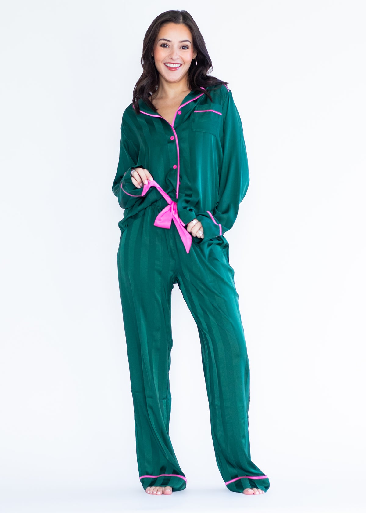 A woman modeling a two-piece green pajama set with bright pink trim and a matching pink tie belt, photographed against a plain white studio background.