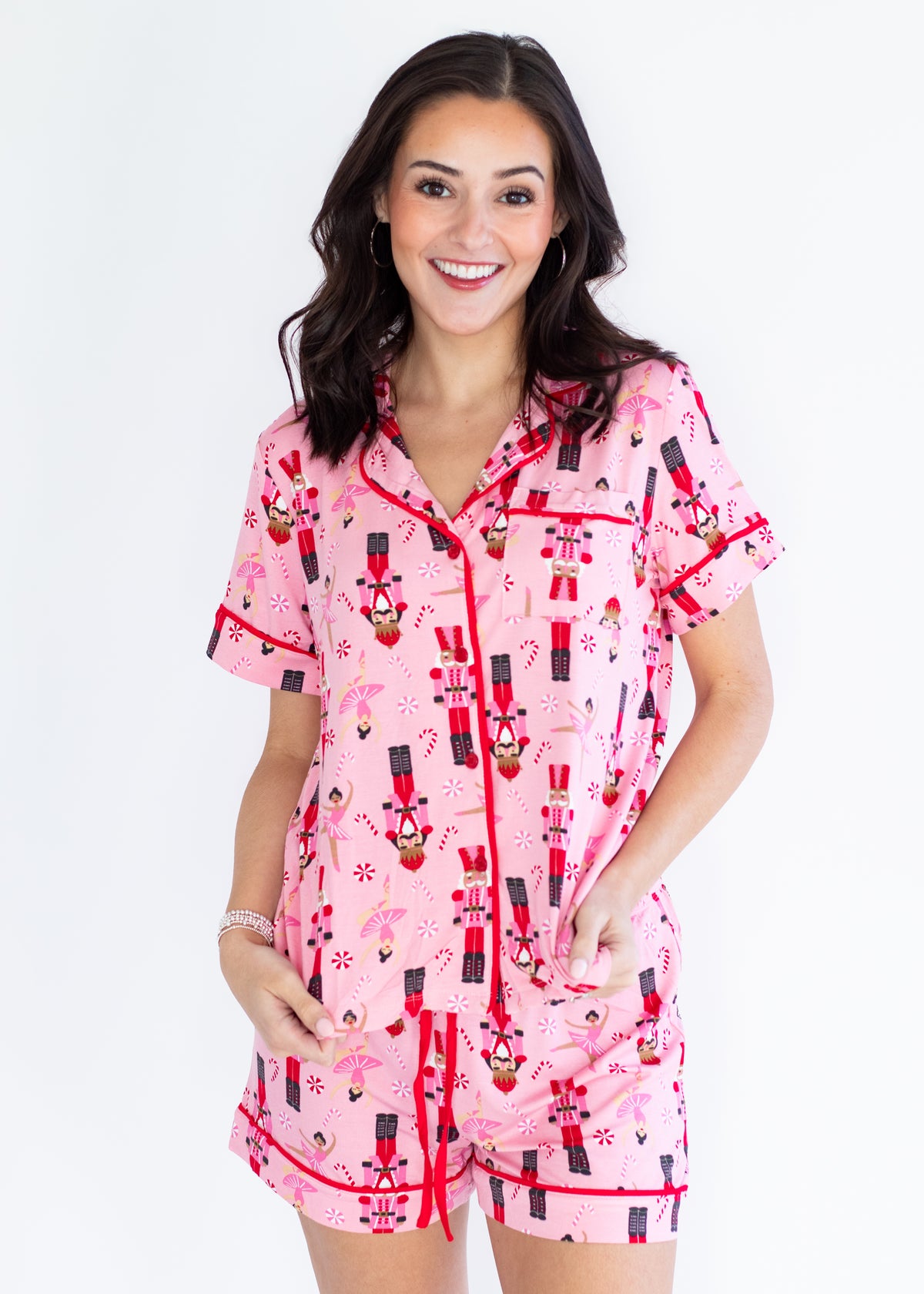A woman modeling a pink short-sleeve pajama set with a nutcracker pattern and red trim, photographed against a plain white background.