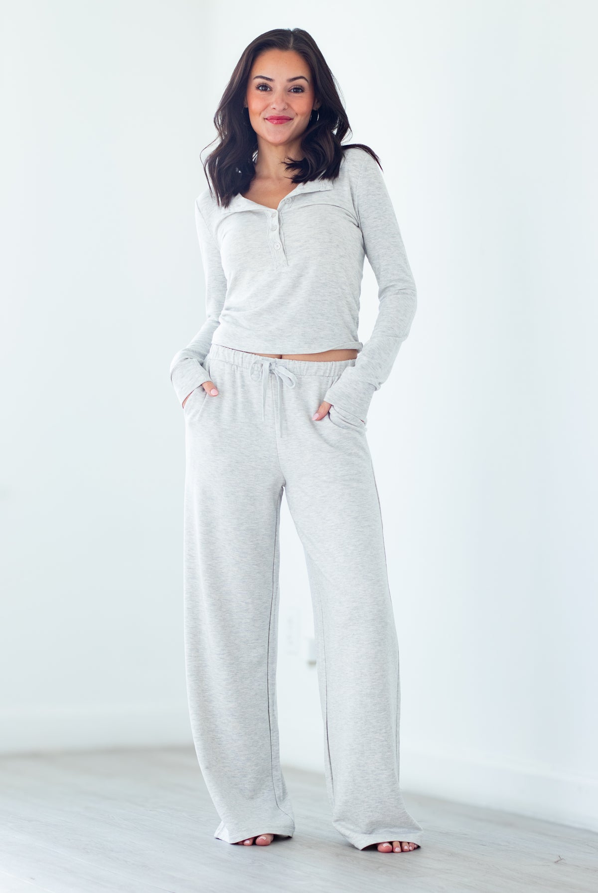A woman modeling a light heather gray lounge set consisting of a long-sleeve half-zip top and matching wide-leg sweatpants, standing in a bright, minimalist room.