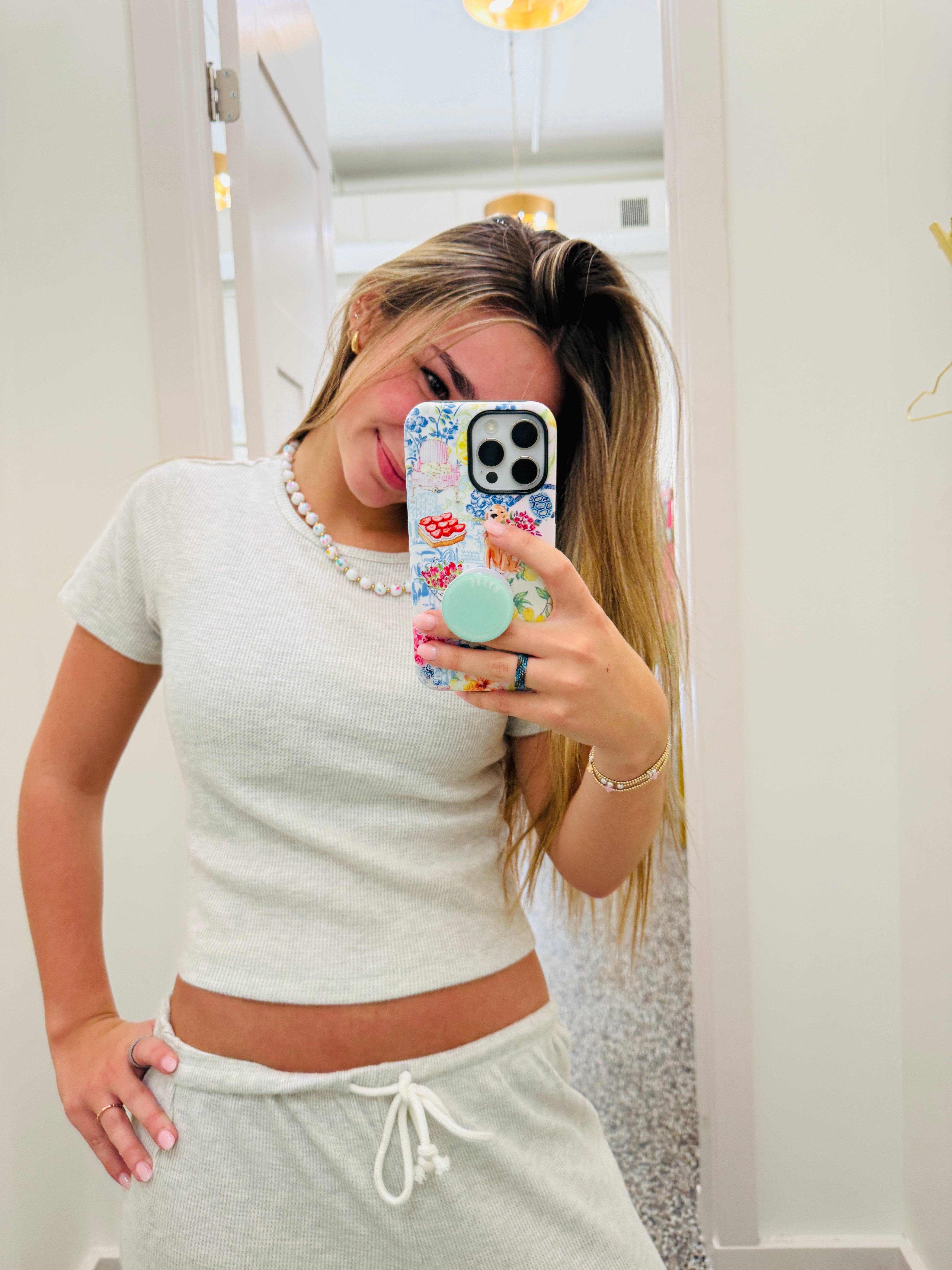A woman taking a mirror selfie wearing a light gray short-sleeve crop top and matching gray sweatpants, holding a floral-patterned phone case with a mint green pop socket.