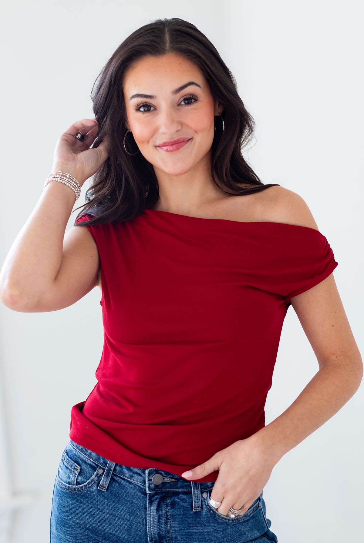 A woman modeling a red off-the-shoulder short-sleeve top with blue jeans, posing against a clean white background.