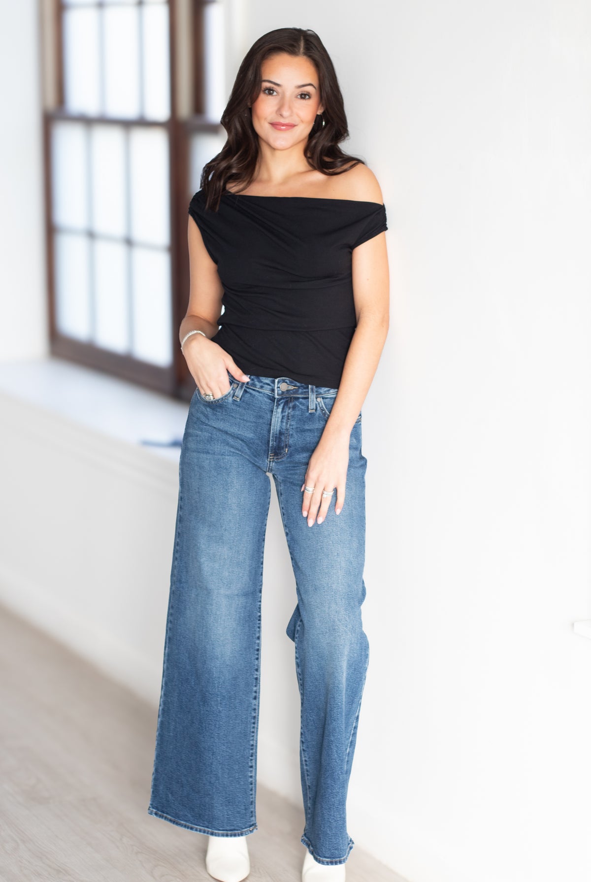 A woman modeling a black off-the-shoulder ruched top and high-waisted, wide-leg blue jeans in a bright, modern room with a large window.