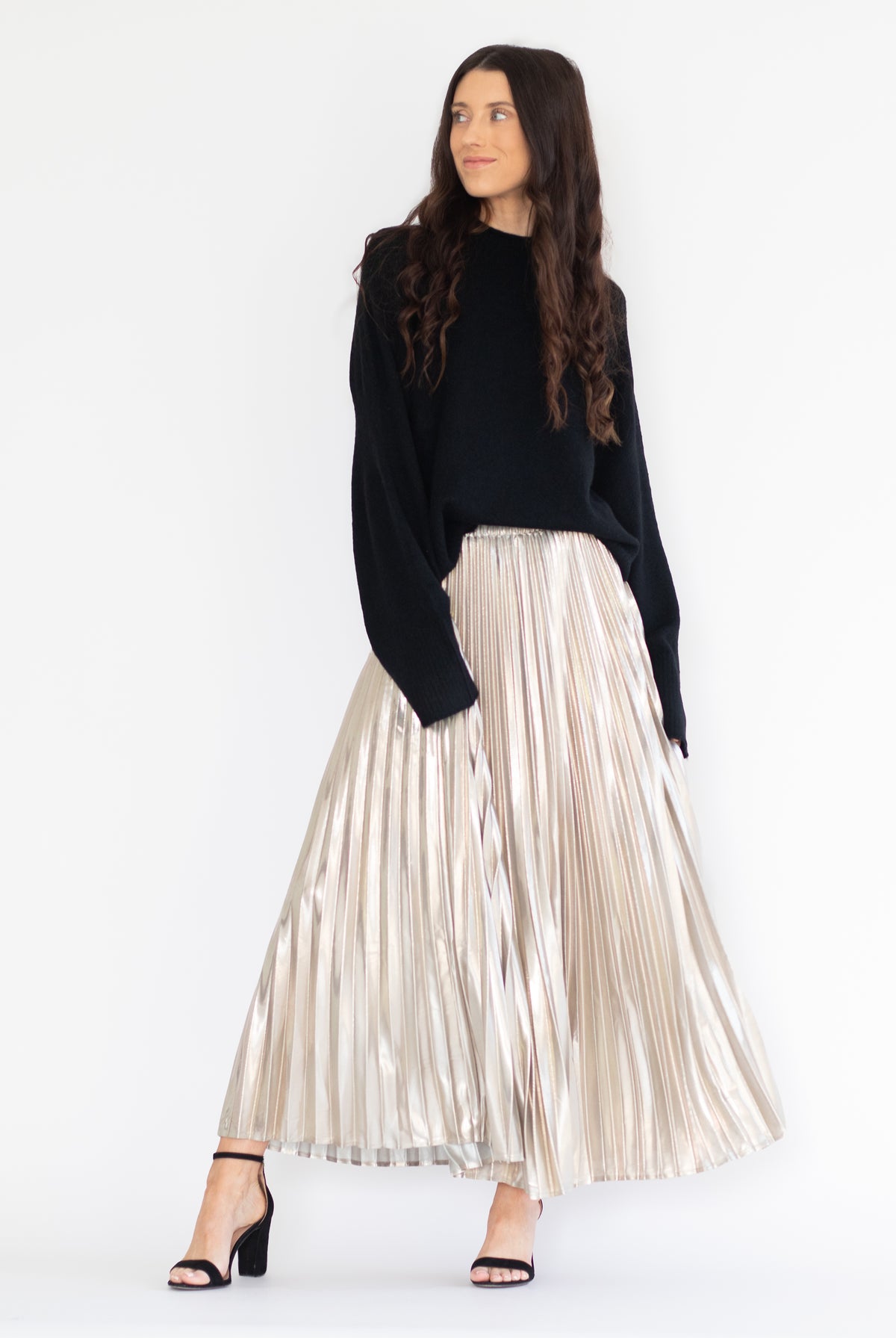 A woman modeling a black knit long-sleeve top and a high-waisted, metallic champagne pleated maxi skirt, paired with black heeled sandals, against a plain white studio background.