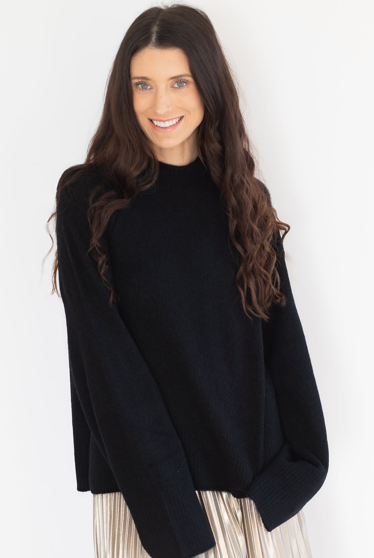 A woman with long, wavy brown hair wearing an oversized black knit mock-neck sweater and a pleated light-colored skirt, posing against a plain white studio background.