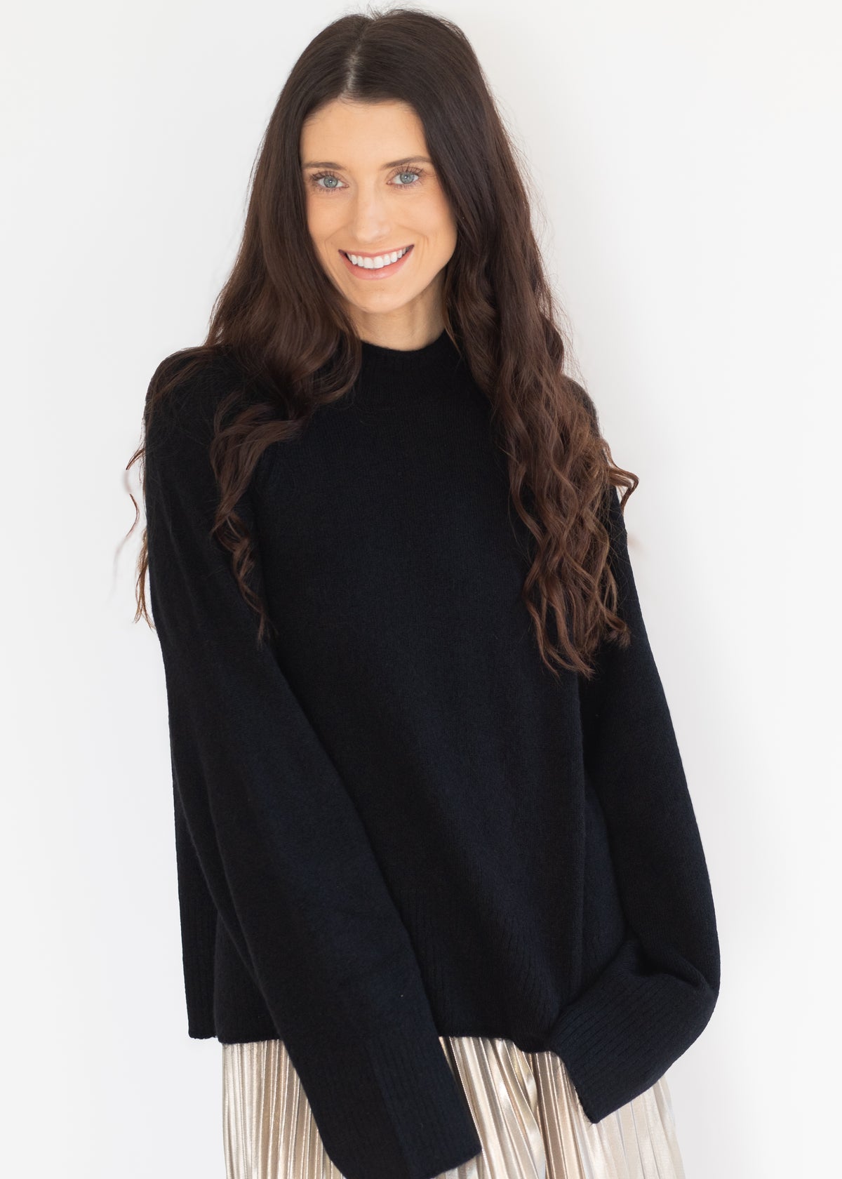 A woman with long, wavy brown hair wearing an oversized black knit mock-neck sweater and a pleated light-colored skirt, posing against a plain white studio background.