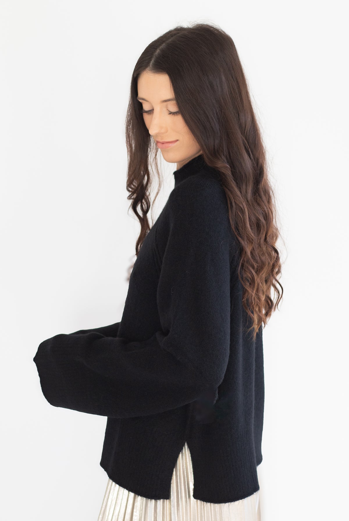 A woman with long, wavy dark brown hair is shown from a side profile, wearing a black knit turtleneck sweater and a pleated cream skirt against a plain white background.