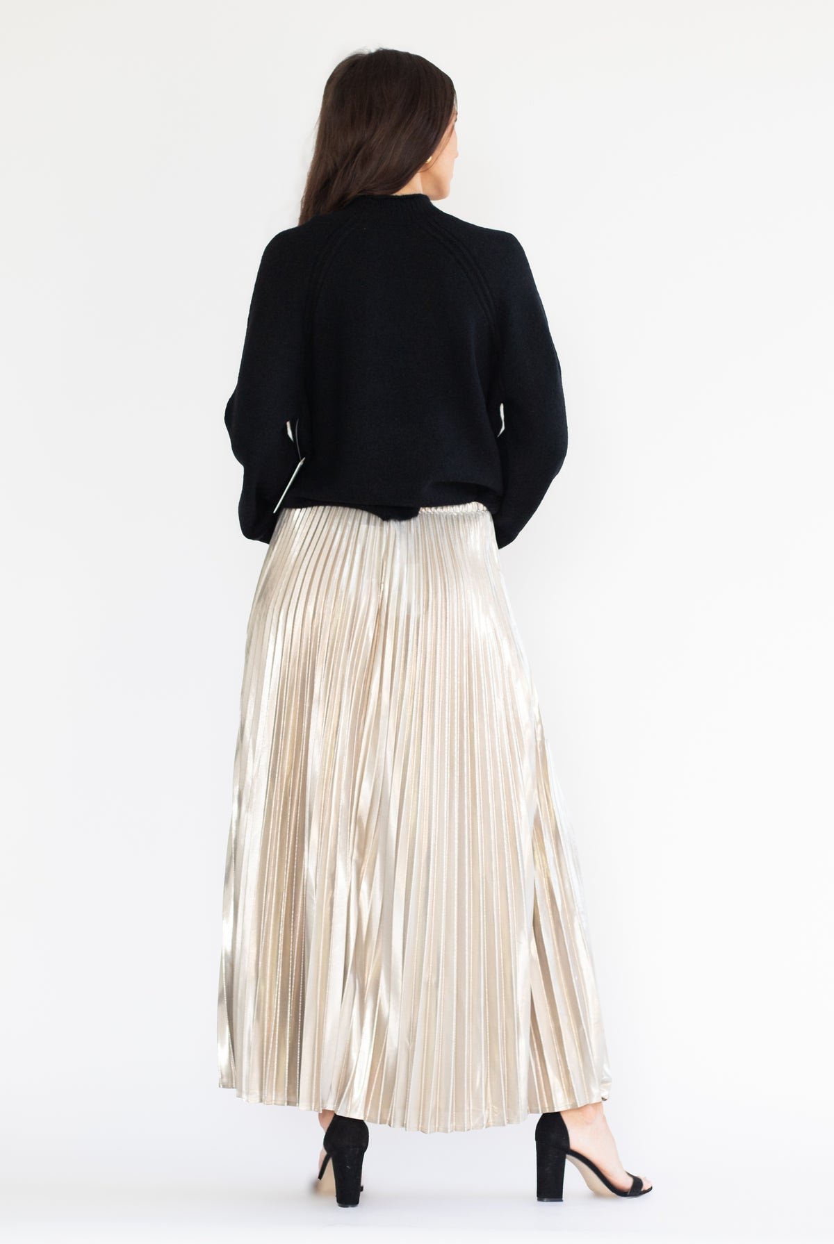 A woman shown from the back wearing a black cropped cardigan and a shiny, pleated champagne-colored maxi skirt, paired with black high-heeled sandals, against a plain white studio background.